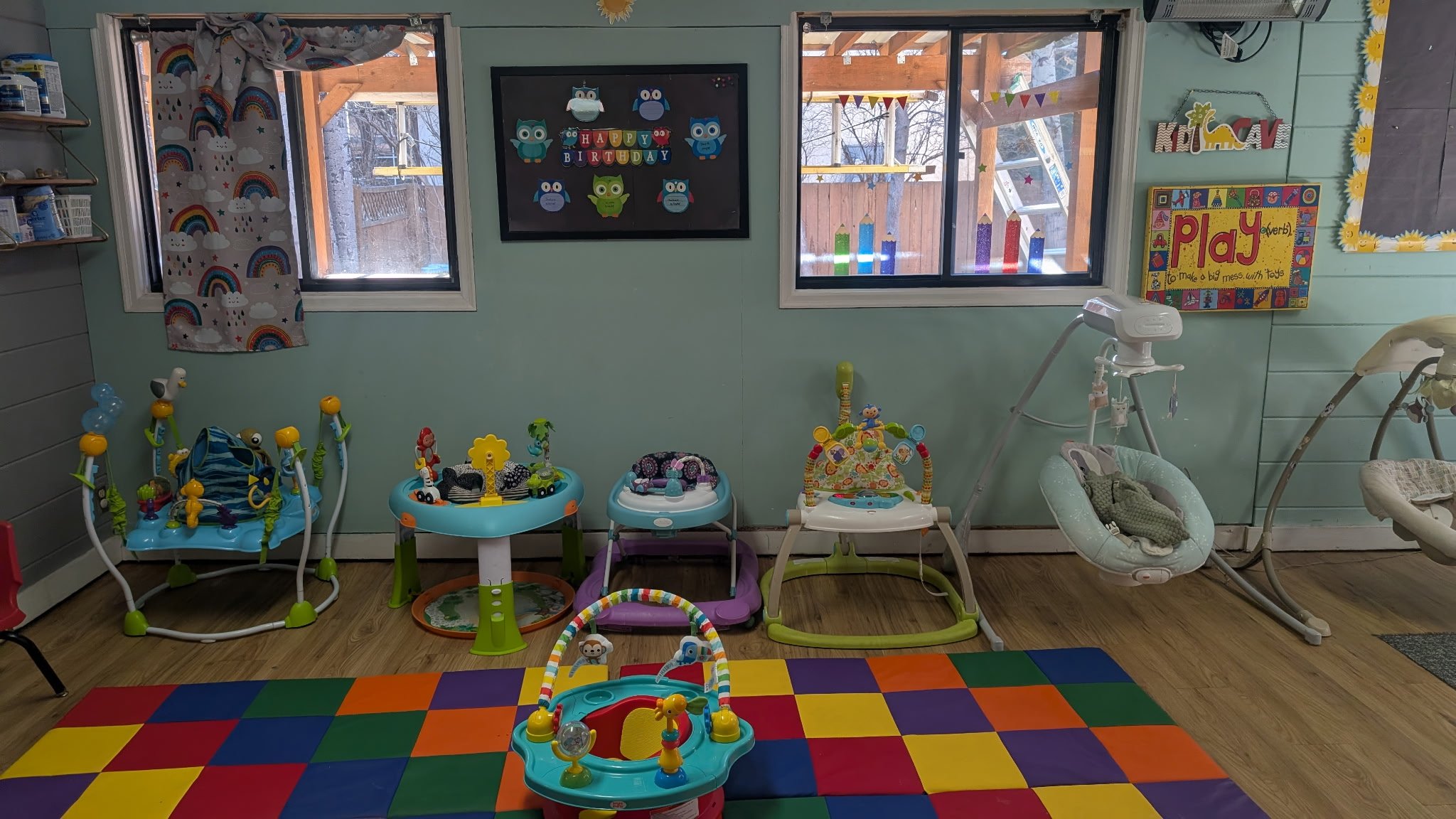 A colorful playroom with large windows, displaying various baby and toddler toys, activity centers, and baby swings, on a multicolored checkered rug.