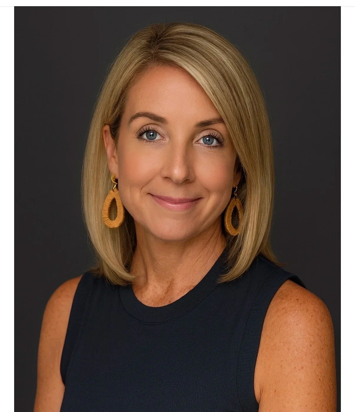 A woman with blonde hair, wearing a sleeveless black top and large, round, woven earrings, smiling against a dark background.
