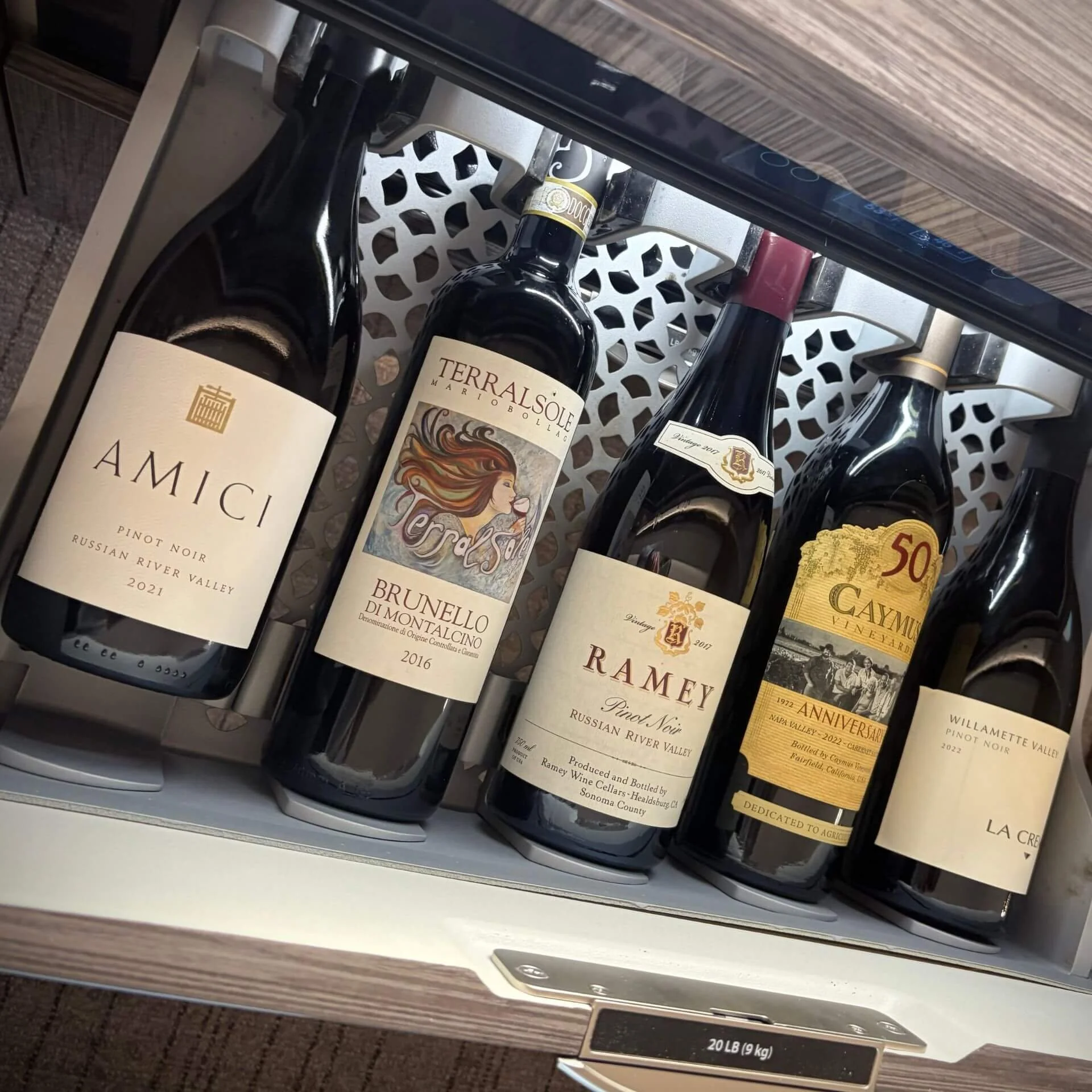 A display shelf with five bottles of wine, including Amici Pinot Noir, TerraSolë Brunello di Montalcino, Ramey Pinot Noir, Caymus Vineyards, and La Crema Pinot Noir.