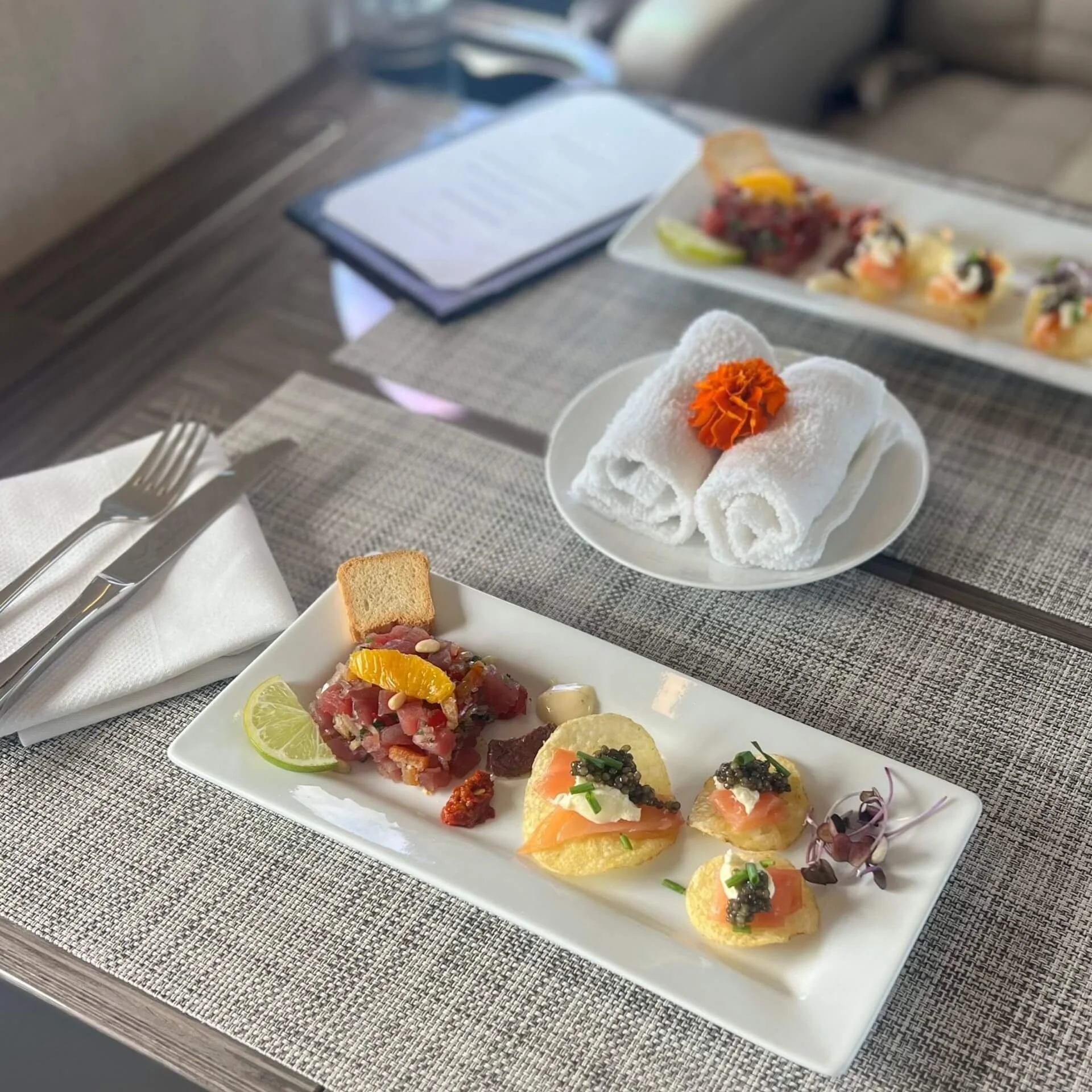 Plate of assorted appetizers including crostini topped with smoked salmon and caviar, and steak tartare with lime and a bread cube, on a white rectangular plate. In the background, rolled towels with an orange flower, another plate of food, a notebook, and cutlery on a woven placemat.