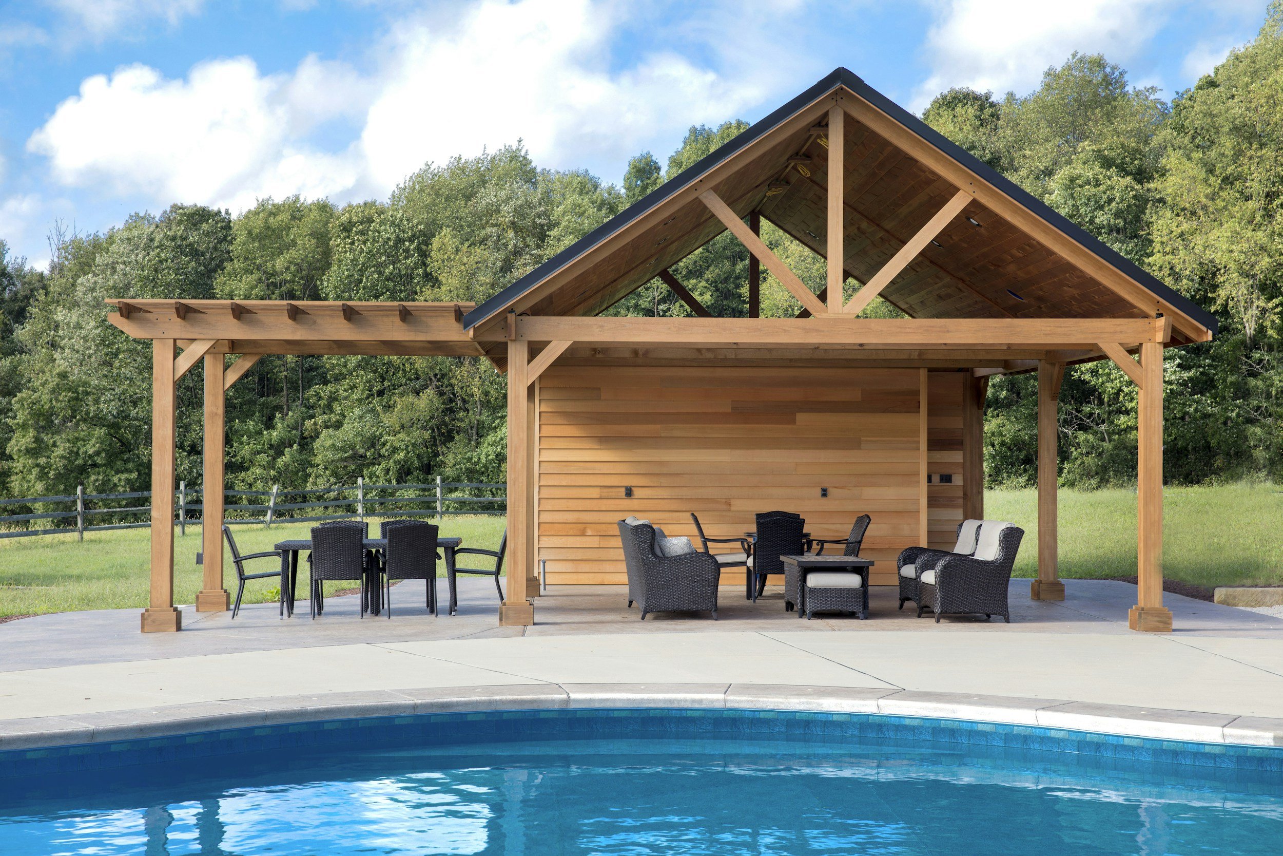 Outdoor poolside area with a wooden covered patio, black wicker furniture, and green trees in the background under a partly cloudy sky.