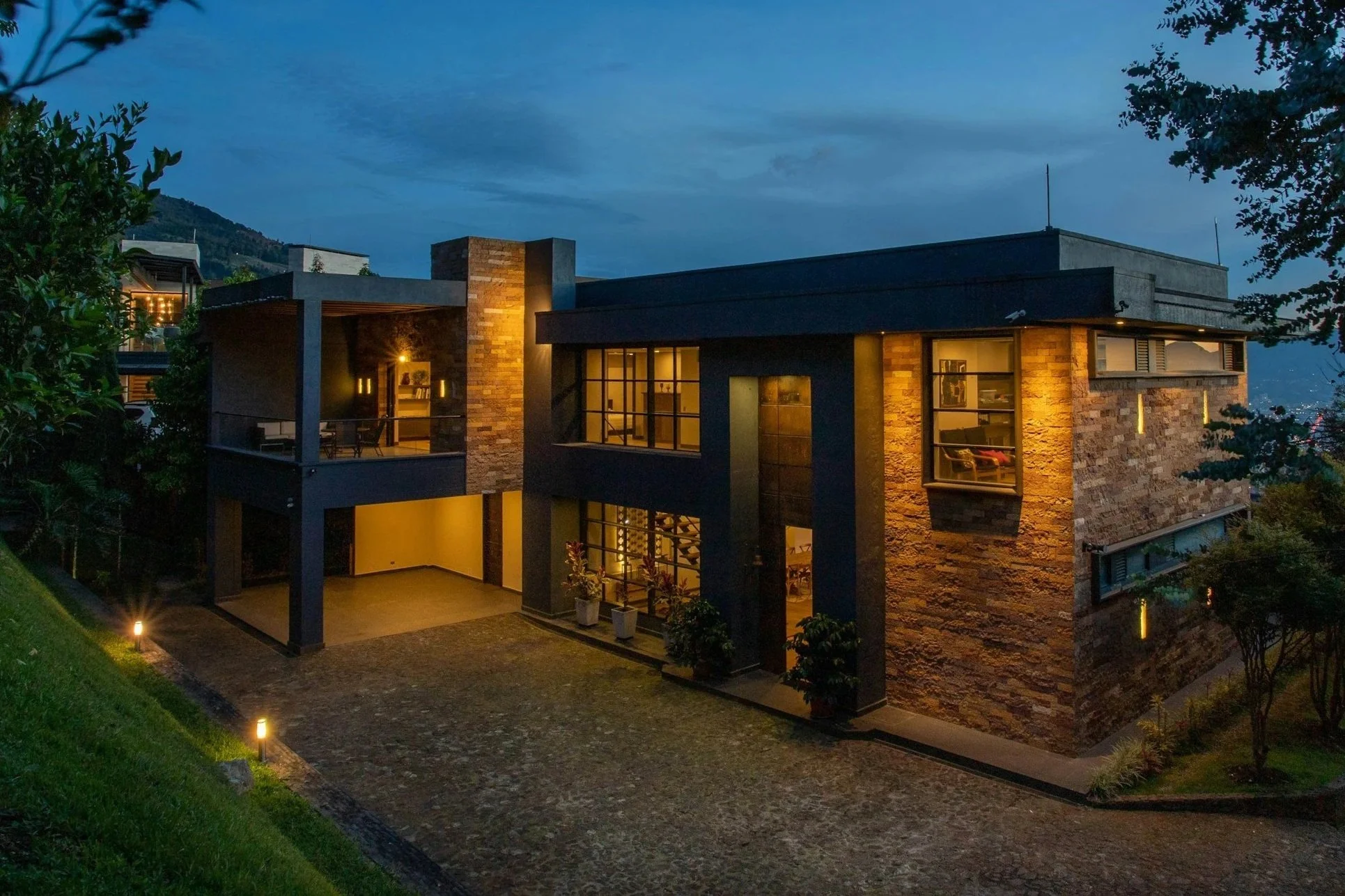 Modern house with stone and dark exterior, illuminated from inside, located on a sloped landscape with trees and a cobblestone driveway, under a cloudy evening sky.