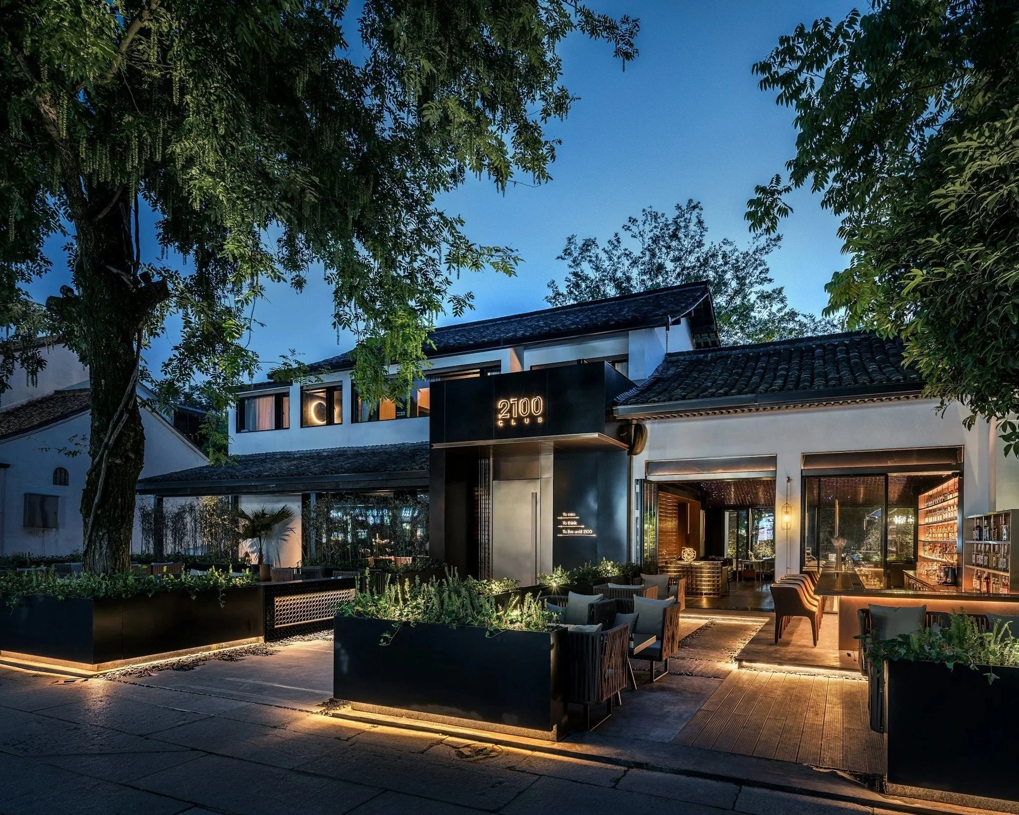 Exterior view of a modern upscale nightclub called 2100 Club, with black signage, outdoor seating area, and large glass windows, illuminated during twilight.