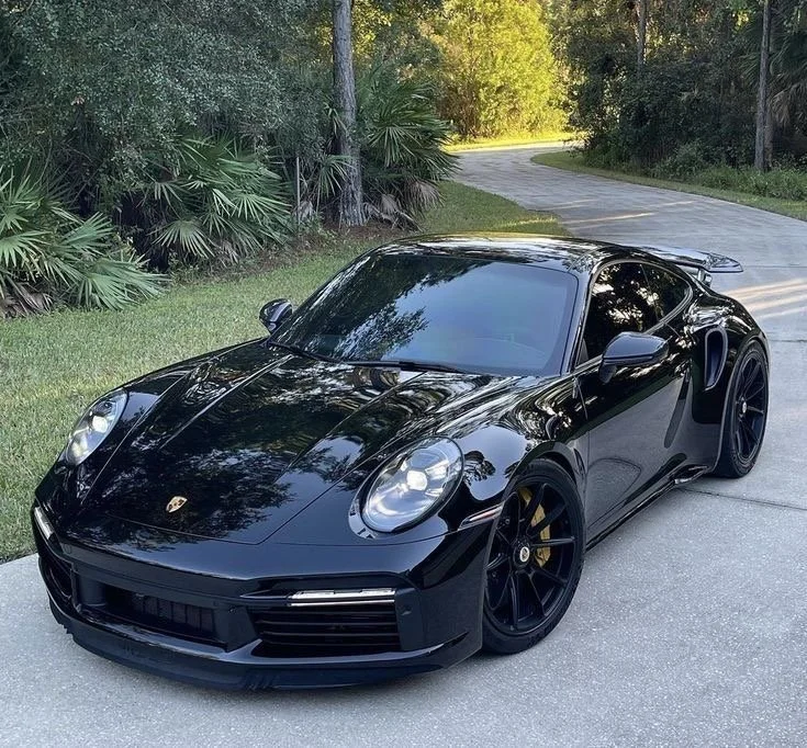 A sleek black Porsche sports car parked on a quiet road with trees and greenery in the background.