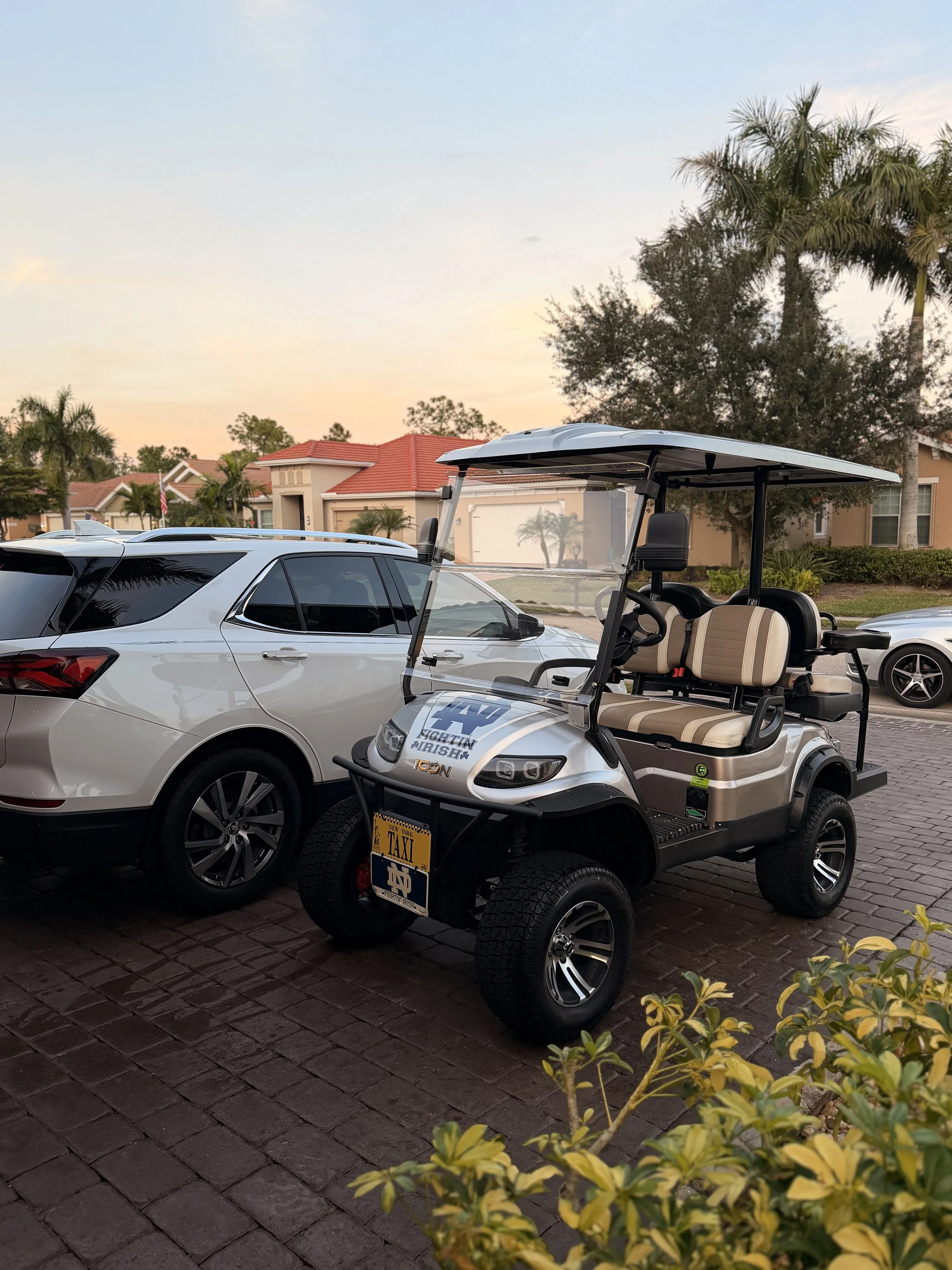 A golf cart parked next to a white SUV on a brick driveway, with residential houses, palm trees, and a sunset sky in the background.
