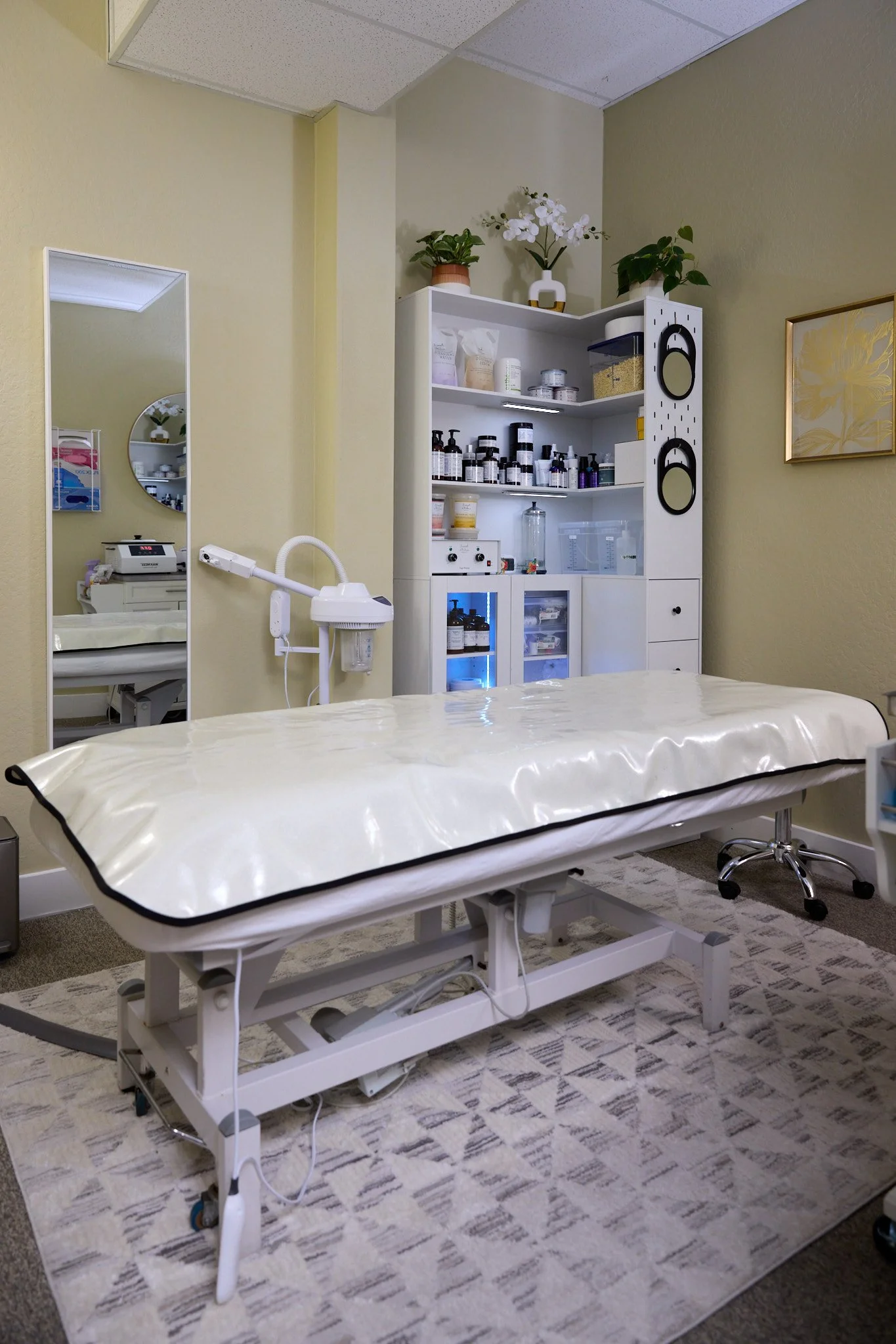 A spa treatment room with a bed, a tall white cabinet with hair removal supplies and equipment, a wall-mounted lamp, a mirror, and decorative plants. 