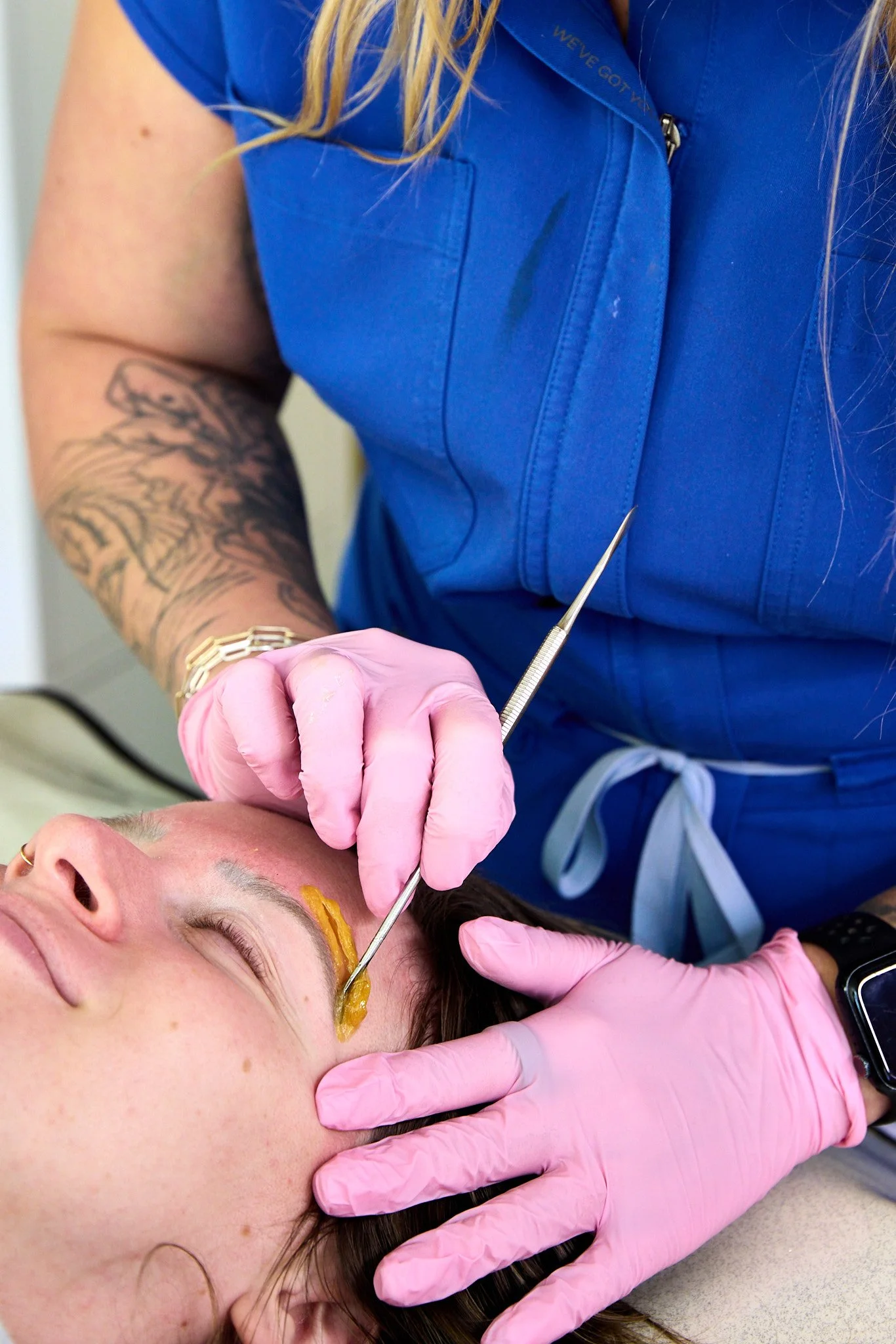 A person receiving an eyebrow sugaring from Rachel, who's wearing pink gloves and a blue uniform.