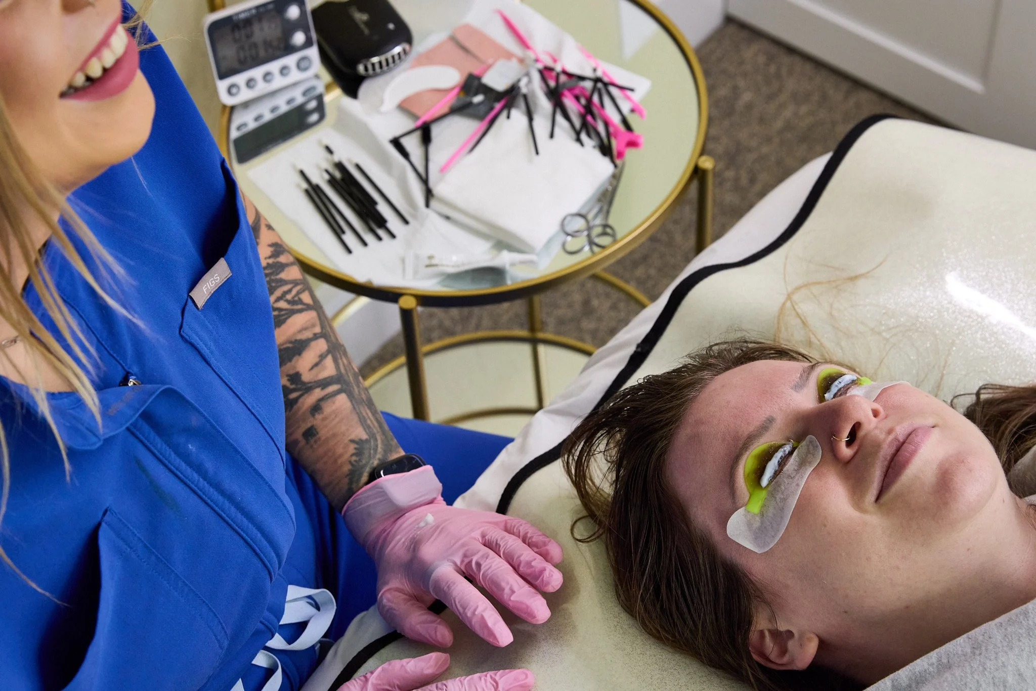 A client lying on a treatment bed while receiving a lash lift. In the background, a tray holds various supplies.