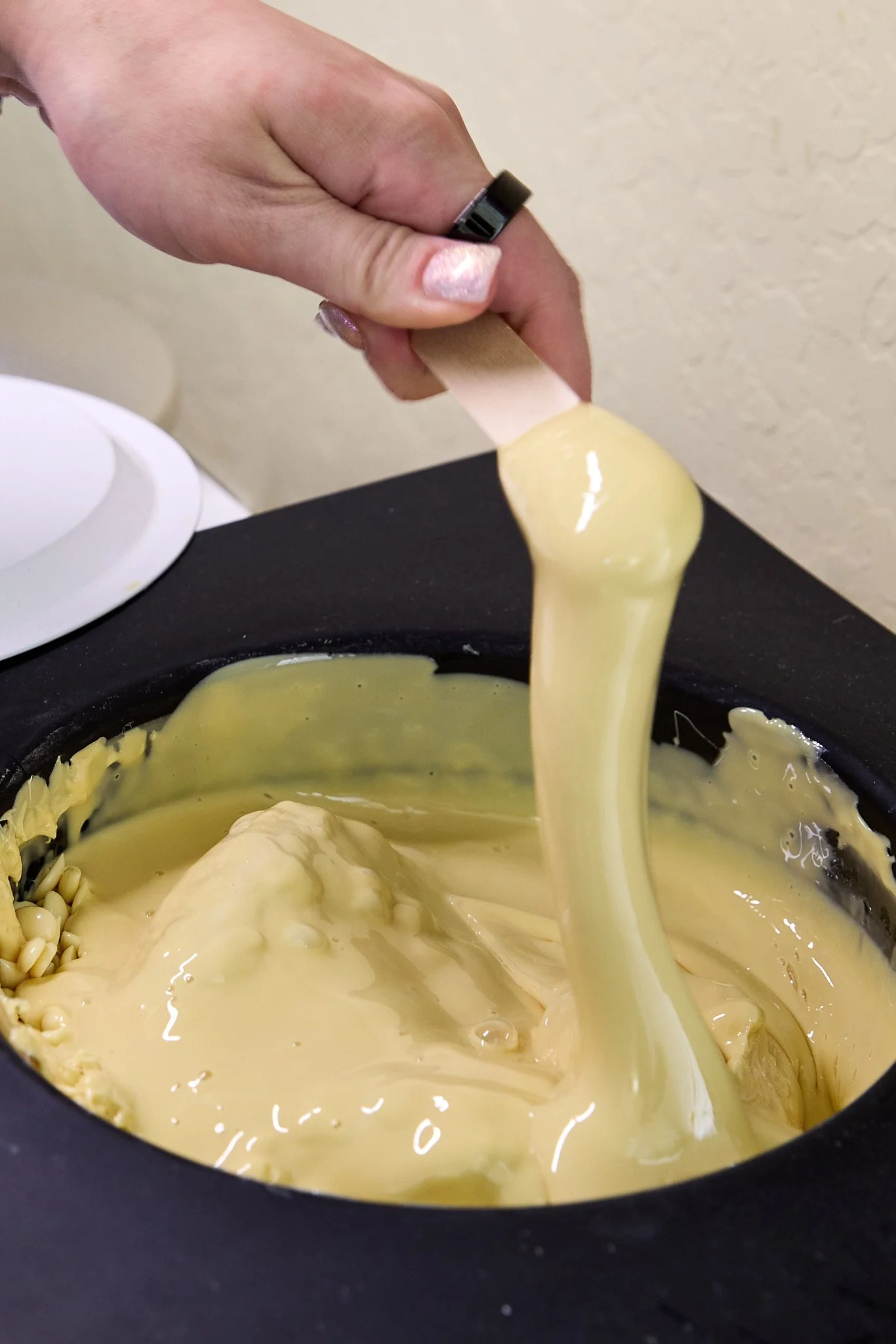 A hand holding a spatula dipping into heated wax.