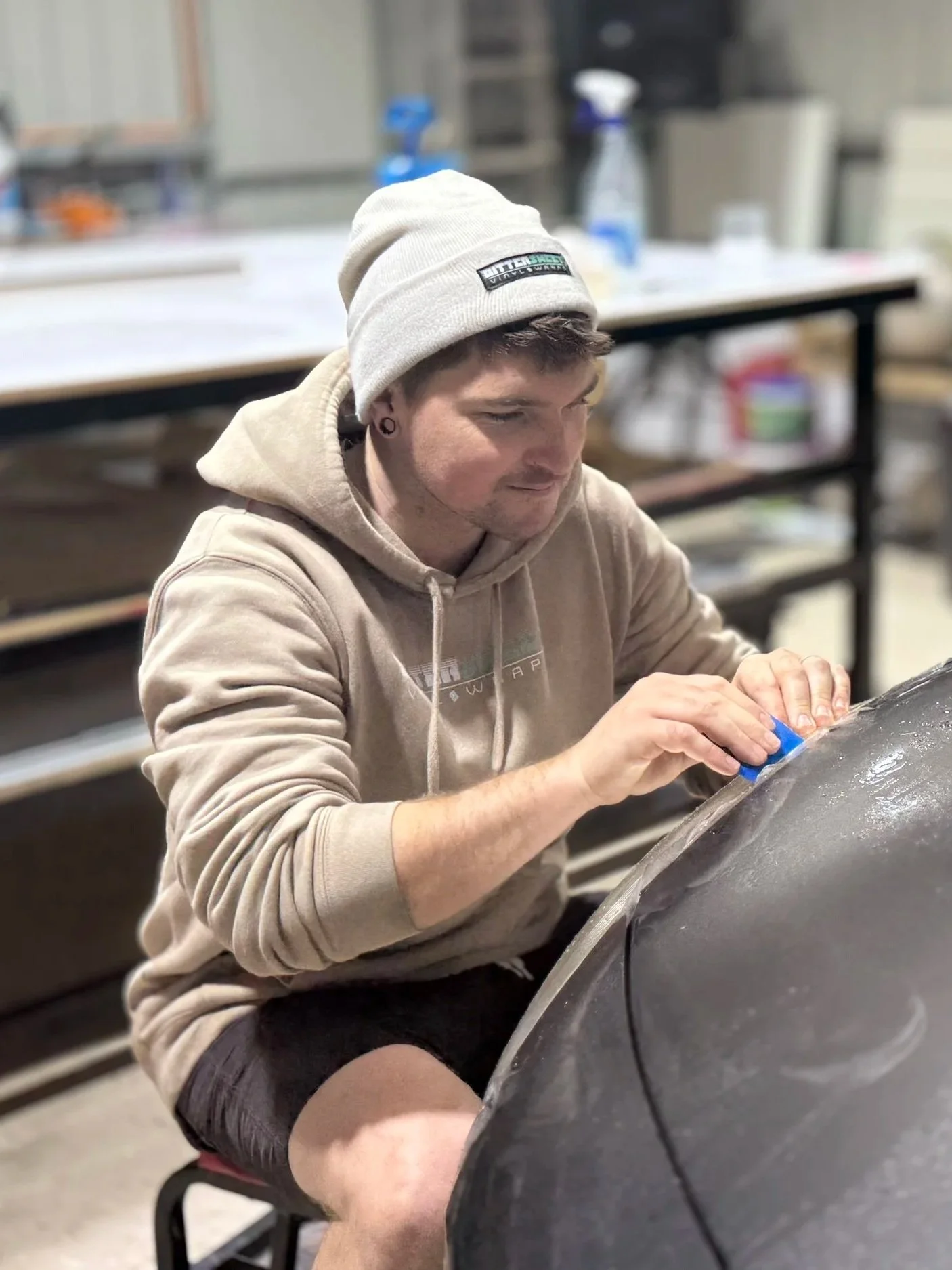 A young man with a beige hoodie and a gray beanie is sanding a large, curved object in a workshop, with shelves and tools in the background.