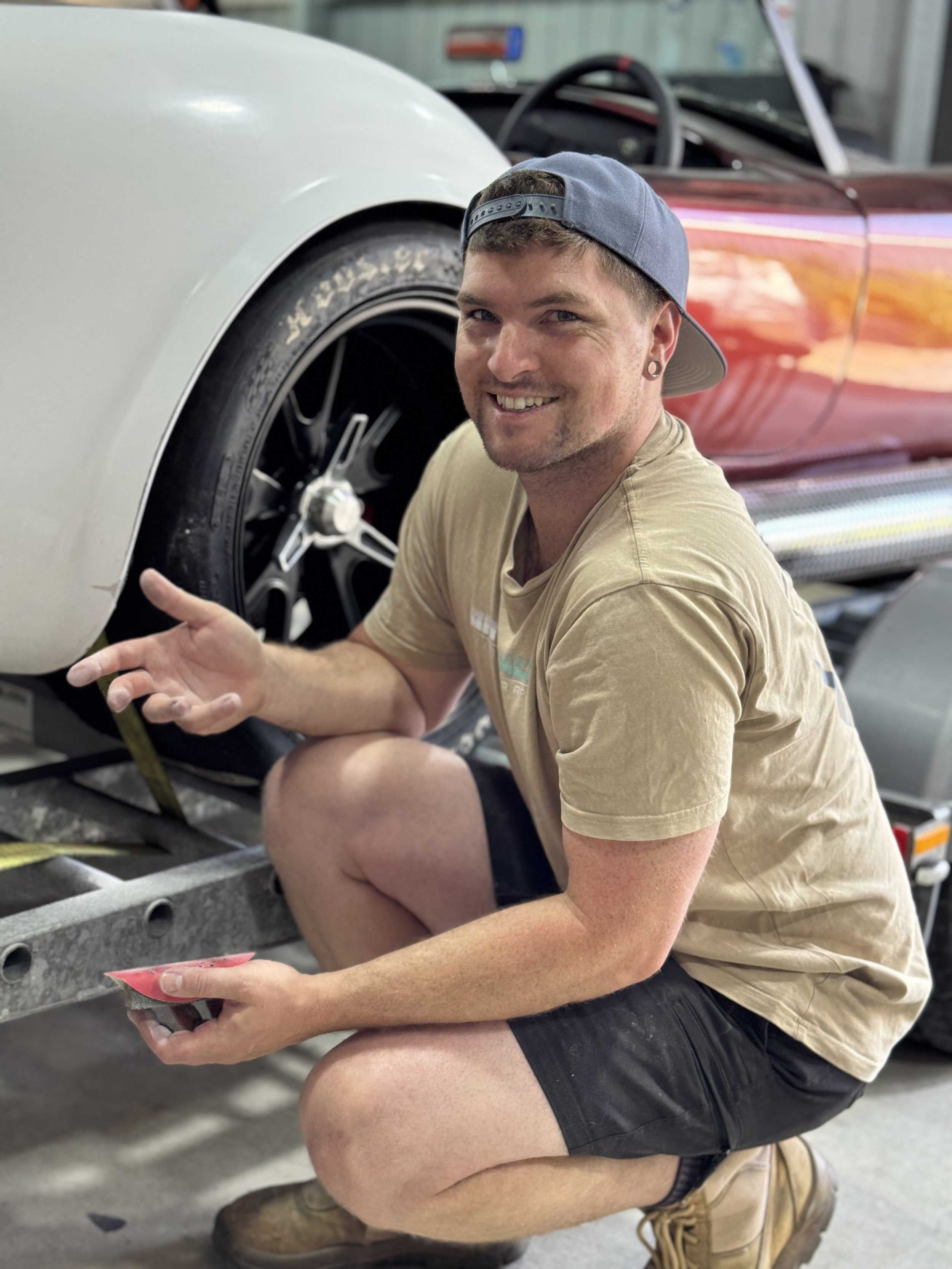A smiling man wearing a backwards baseball cap, tan t-shirt, black shorts, and tan work boots crouches next to a vintage car, holding a small item in his hand. The car is on a trailer or platform with part of the wheel and body visible, and the background shows a garage or workshop setting.
