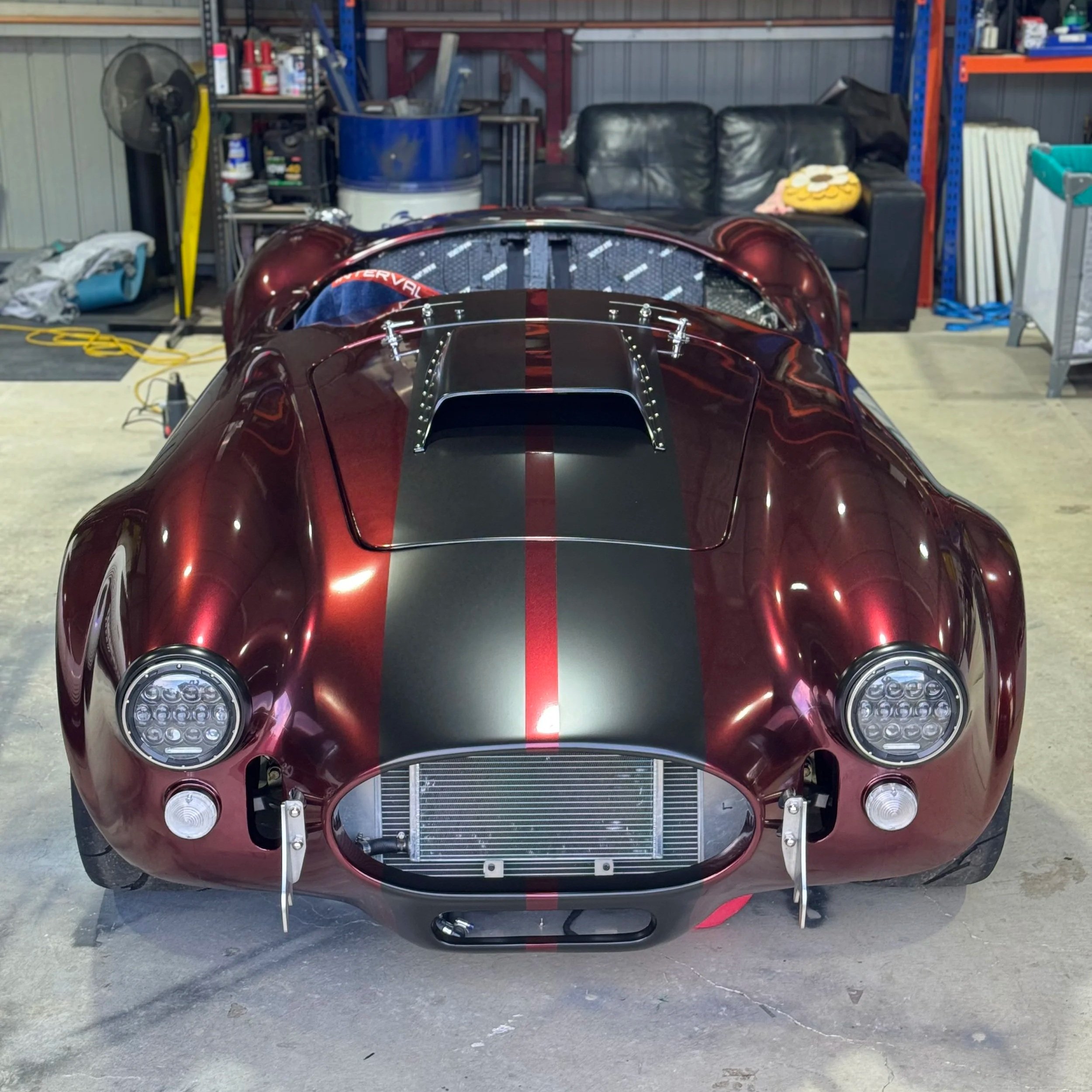 A vintage race car with a sleek, red paint job, black racing stripe down the center, and a partially exposed engine and cooling radiator in a garage or workshop.