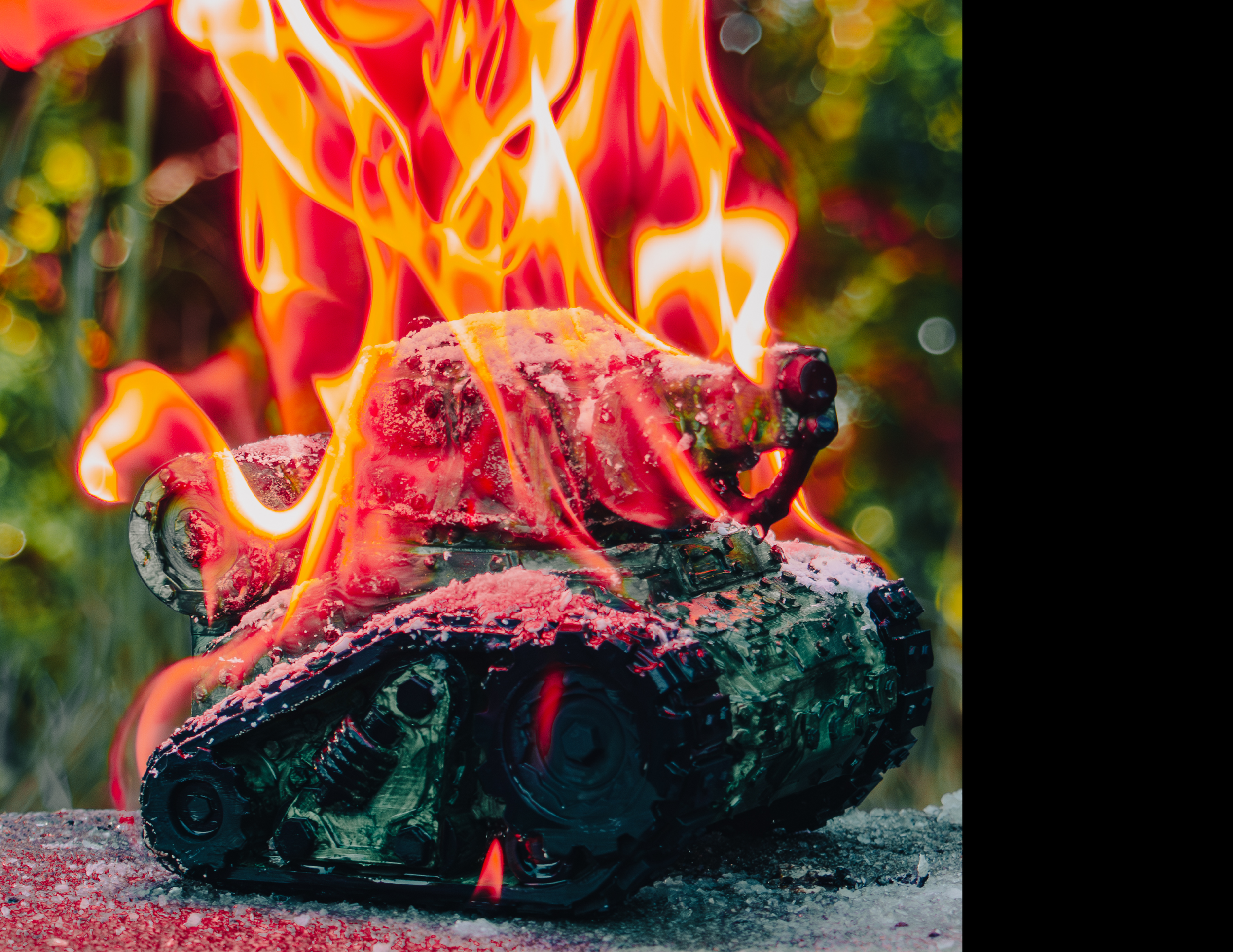 A burning tank scultpure with red flames, ashes, and melted parts, on a surface with ash, with a blurred background of greenery.