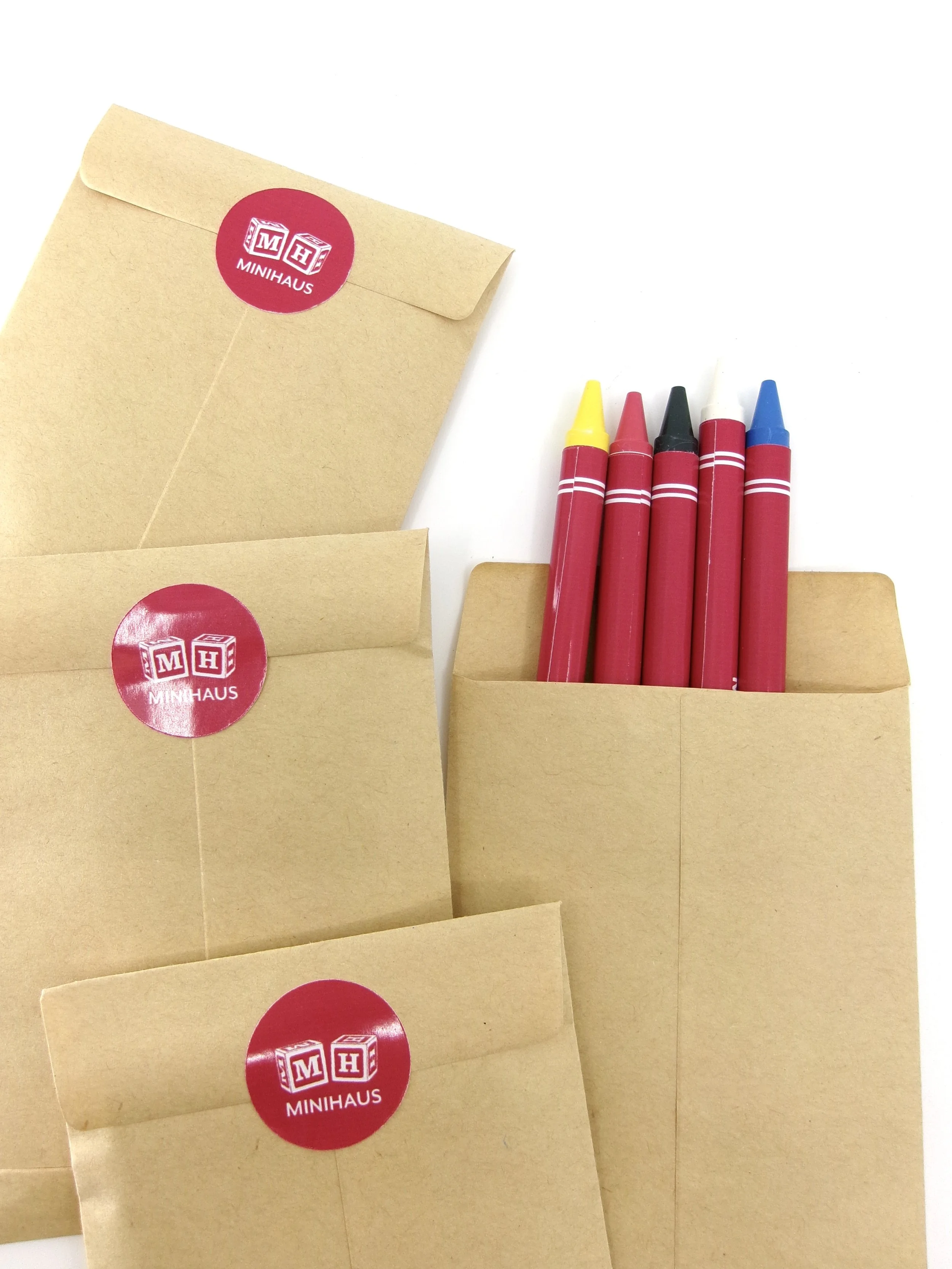 Three brown paper envelopes with red stickers labeled 'MINIHAUS' and a collection of red and yellow, white, blue, and black crayons in one of the envelopes on a white background.