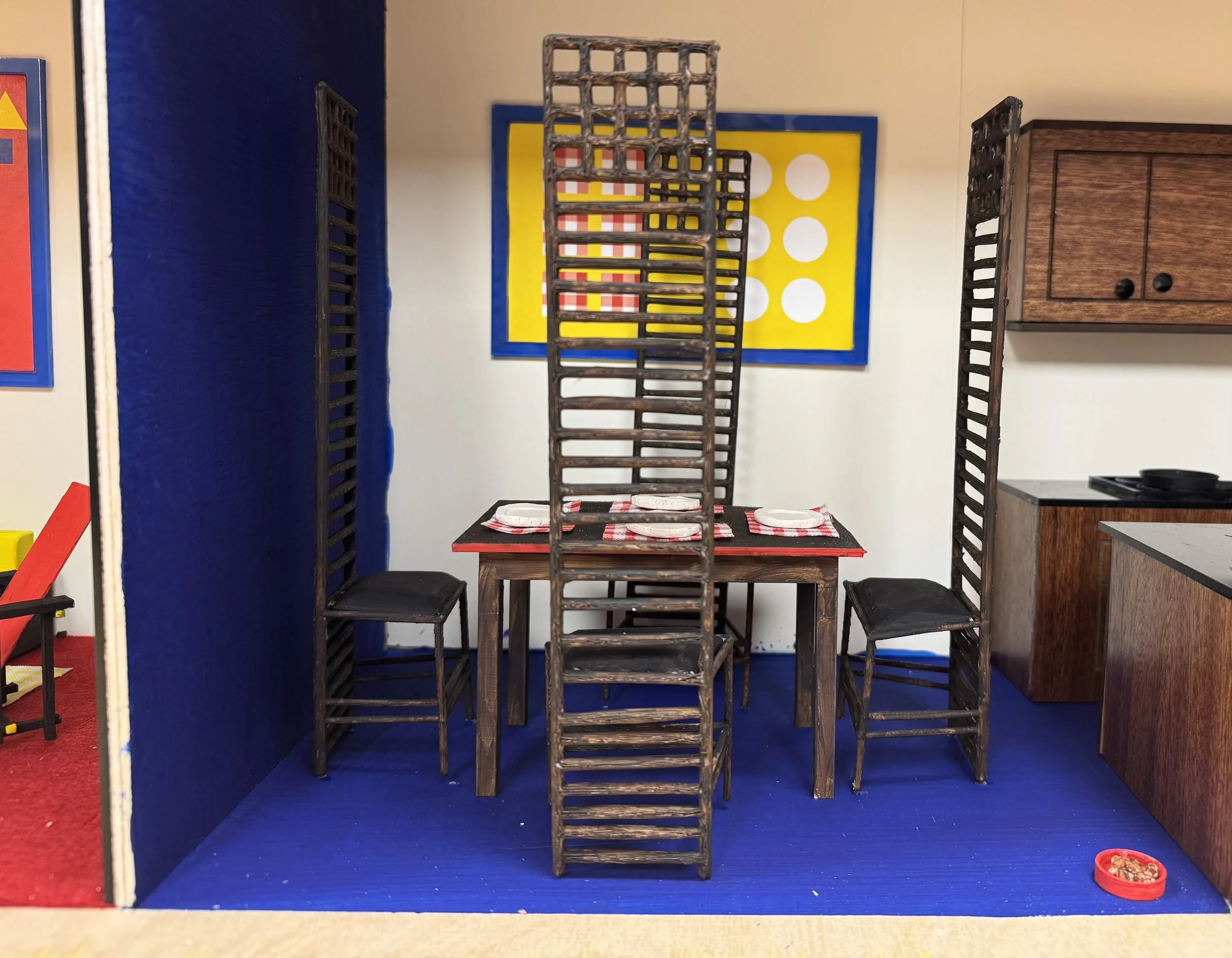 Miniature dining area with a table set for four, surrounded by tall wooden chairs, with a colorful game board on the wall and a small kitchen area to the side.