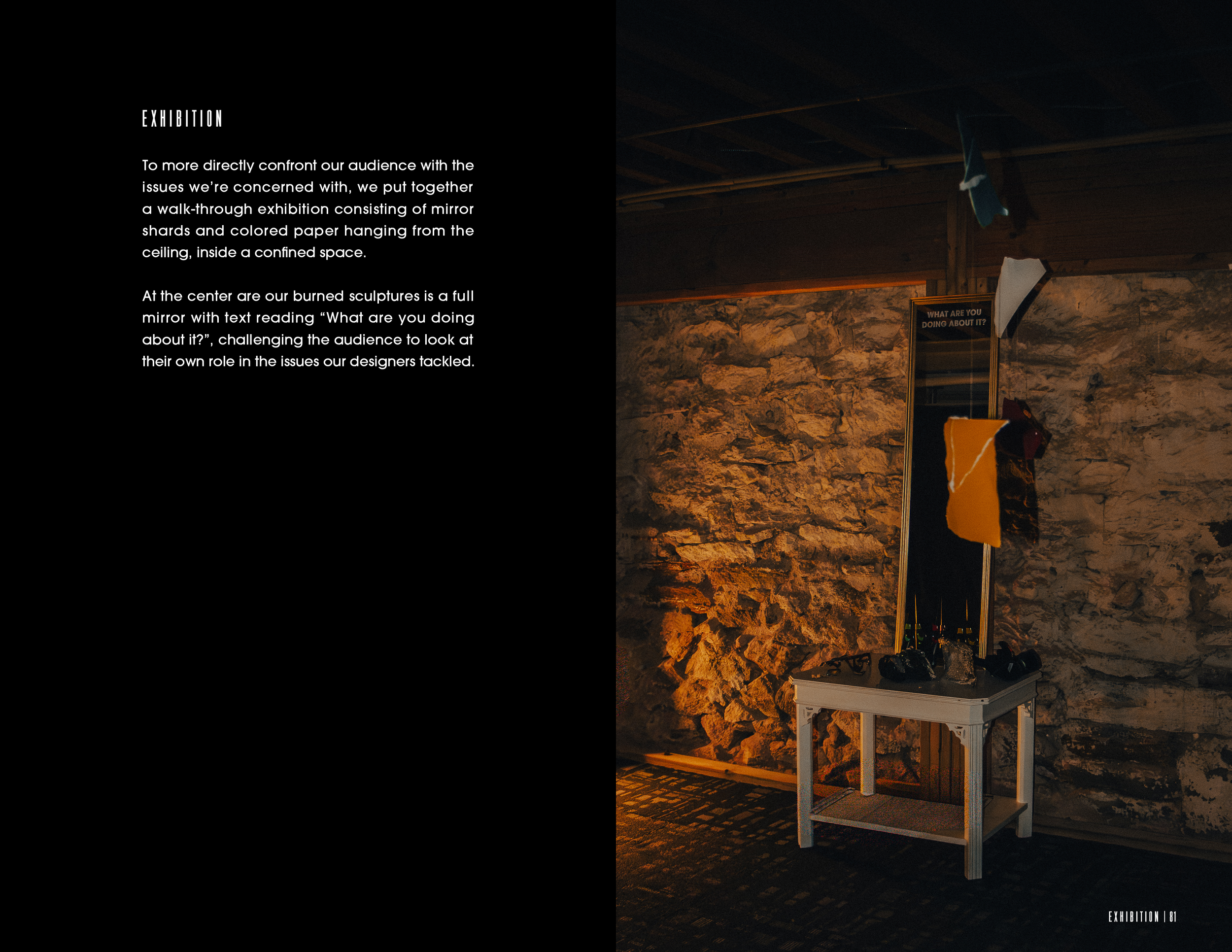 Exhibition display with a black wall on the left containing white text and a corner of a room on the right showing a stone wall, a mirror with text reading 'What are you doing about it?', and clothes hanging above a white table with bottles and other items.