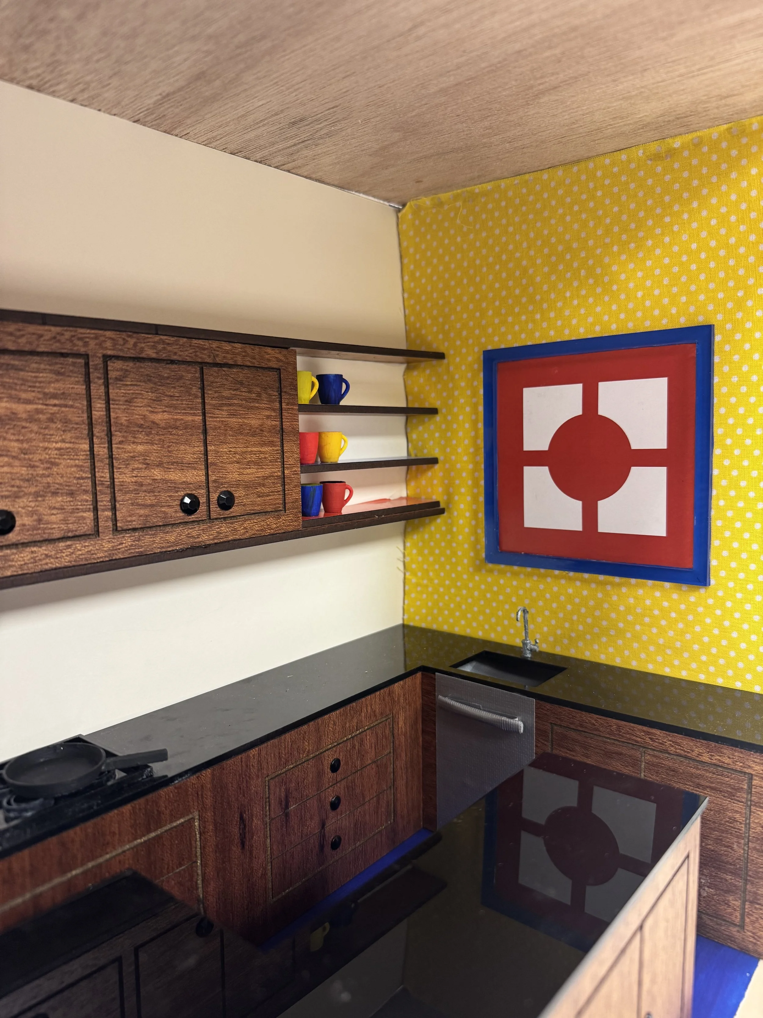 A miniature kitchen model with wooden cabinets, a black countertop, a stainless steel sink, and a backsplash of yellow polka dot wallpaper. There are three small colorful cups on black shelves and a framed artwork with red, white, blue, and black geometric shapes on the wall.