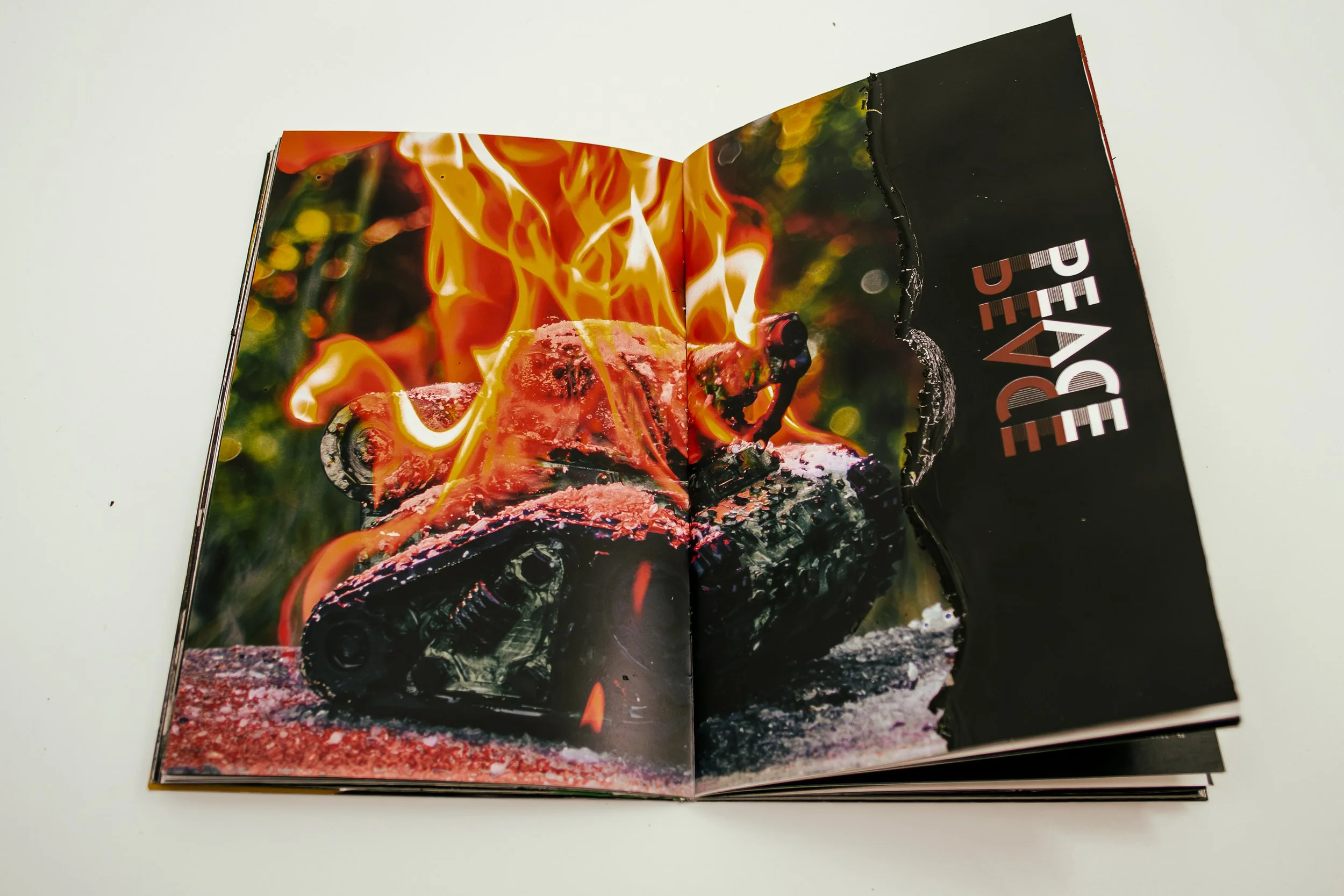 Open magazine featuring a photo of a burning sculpture of a tank with flames and smoke, and text reading 'PEACE' partially visible.