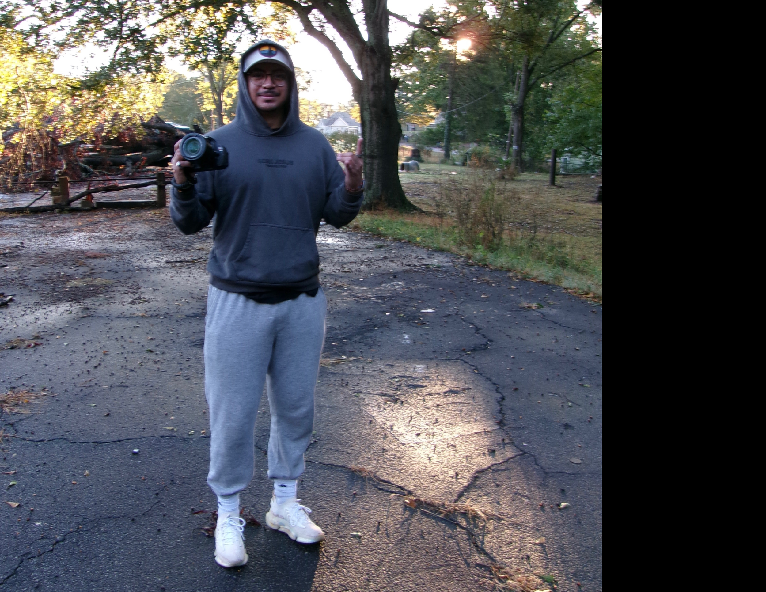 A man standing outdoors on a cracked asphalt surface, holding a camera, with trees and damage from a fallen tree in the background, during what appears to be late afternoon or early evening.