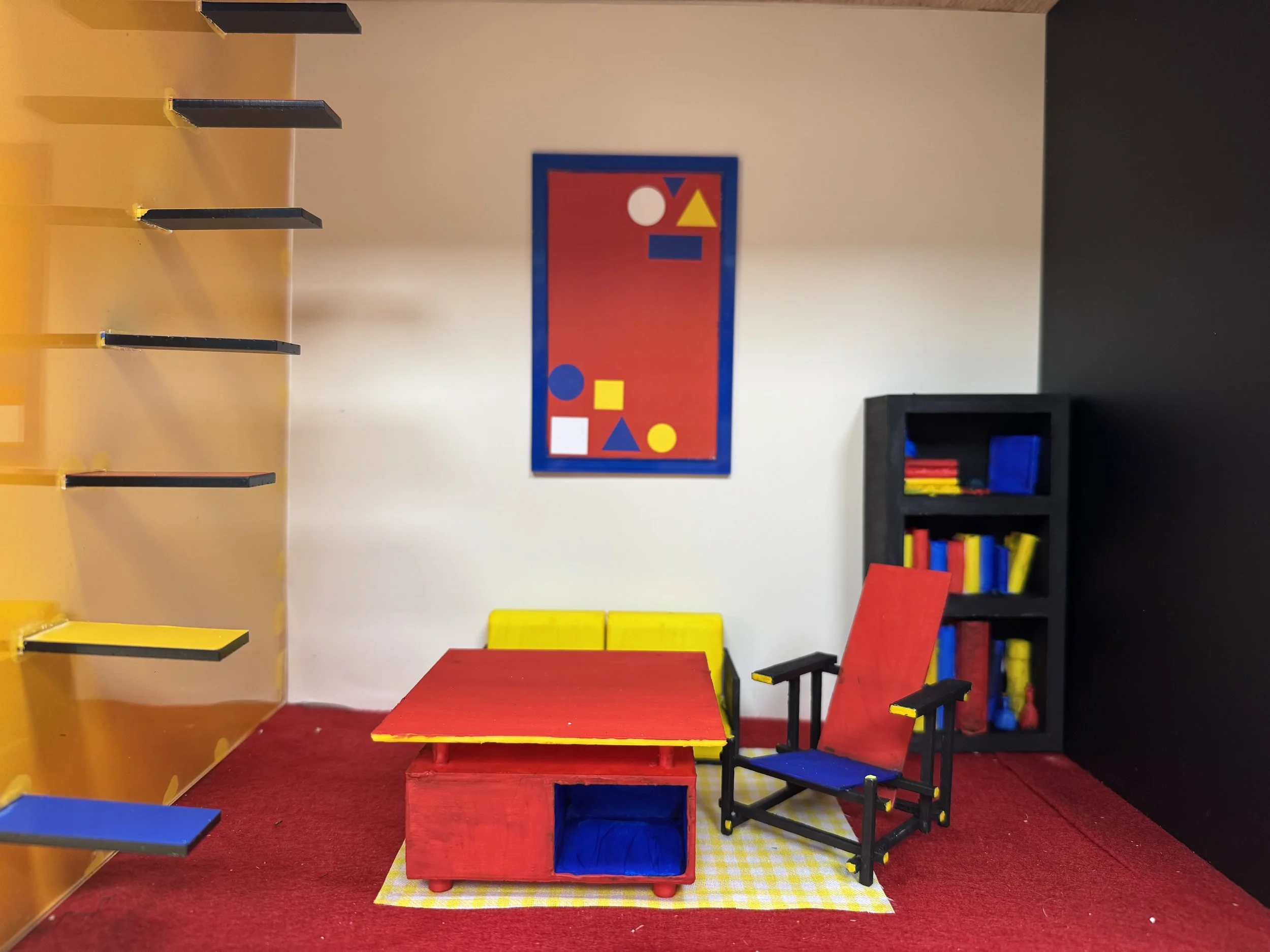 Miniature room with a red table, a blue and red chair, a black bookshelf with colorful books, colorful wall art with geometric shapes, and a red and yellow floor mat.