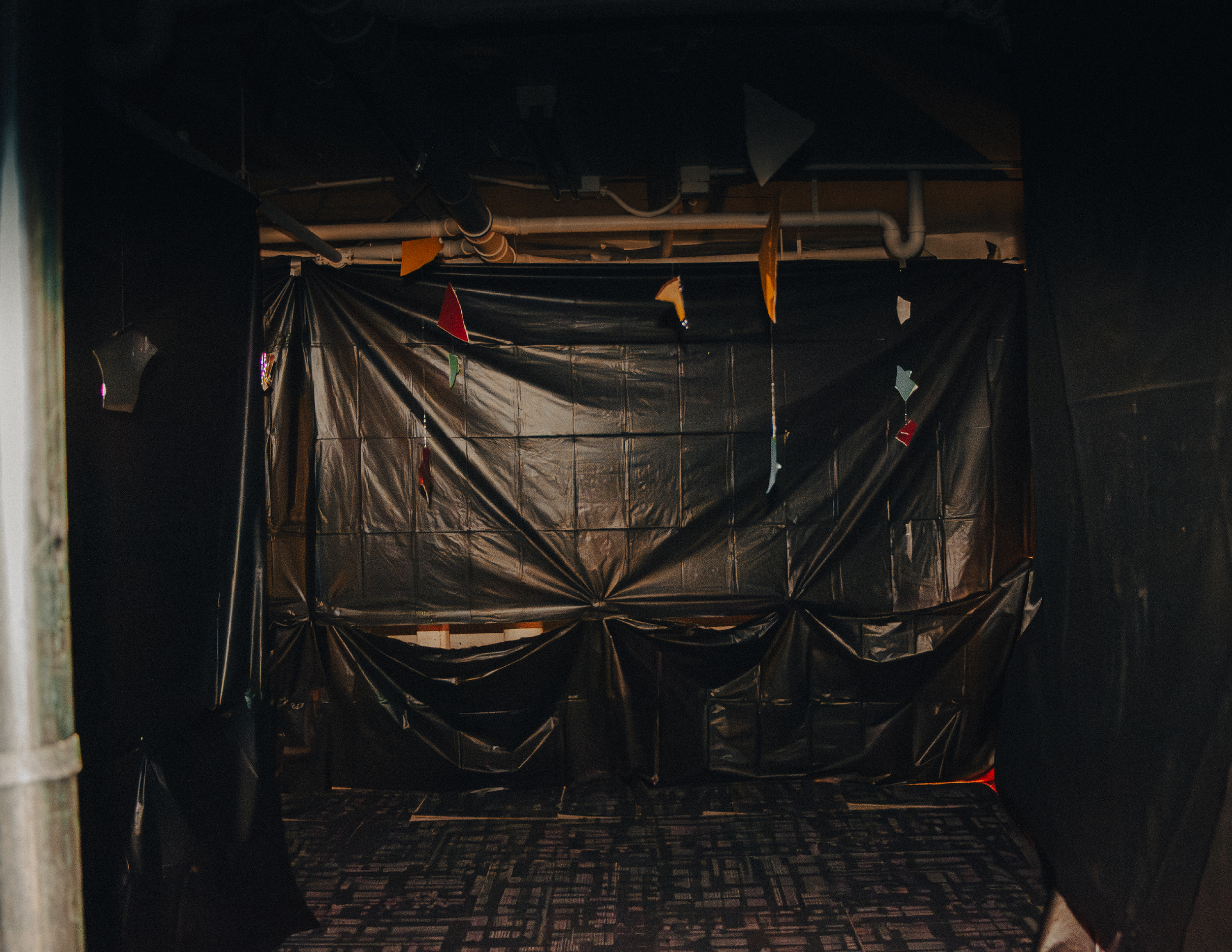 A dark room with black plastic sheeting hanging and covering the walls and ceiling, with colorful paper cutouts and glass shards hanging from a string across the space.