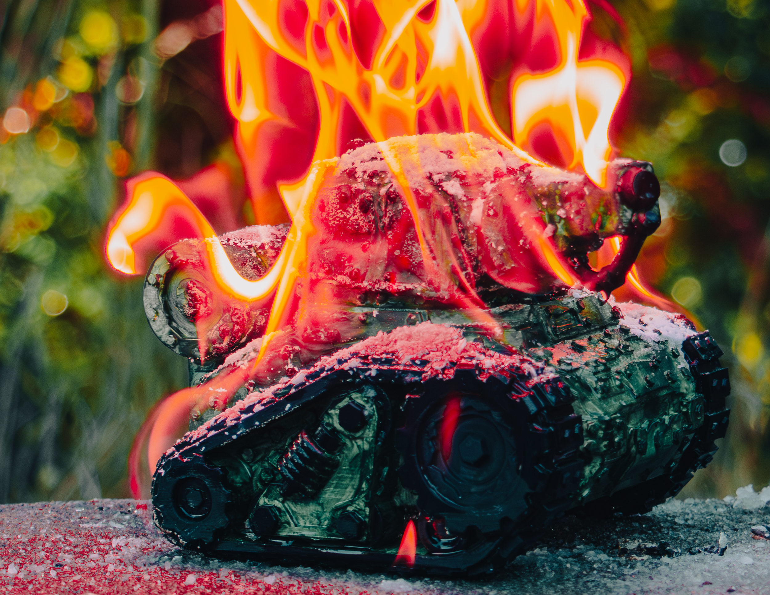 A tank sculpture on fire outdoors with red flames engulfing the vehicle, on a surface with a green blurred background.