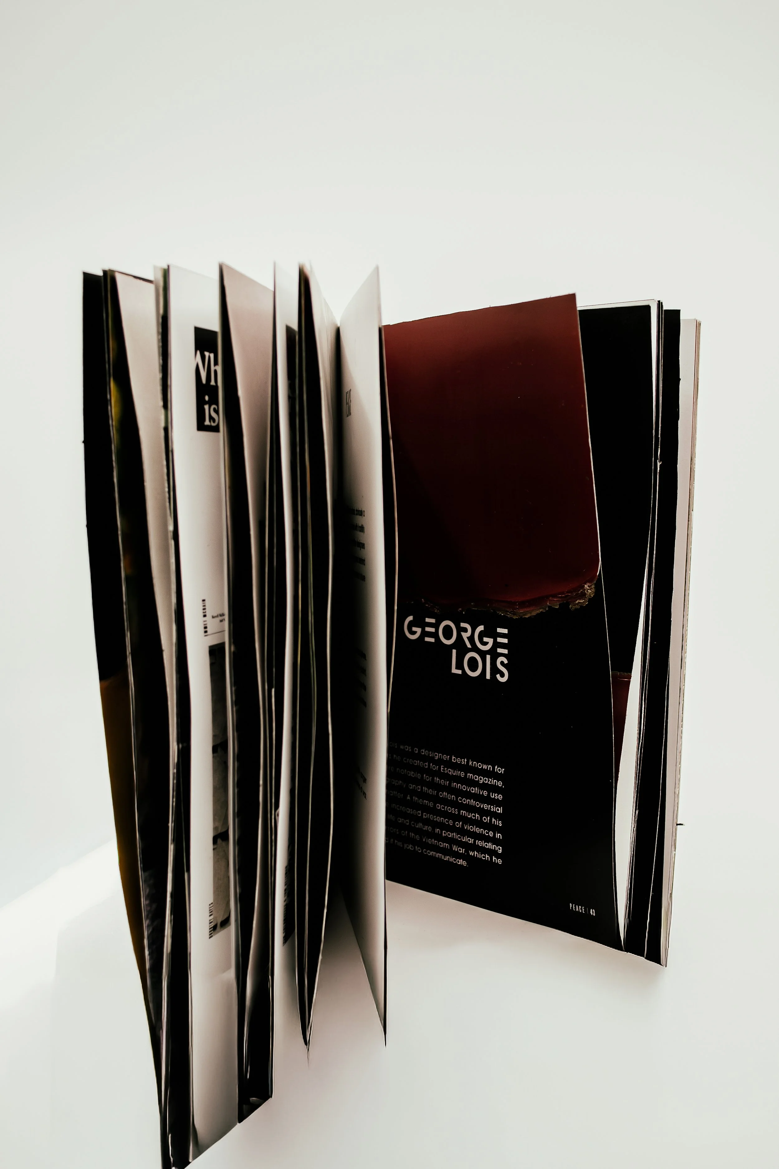 An open magazine placed on a white surface, showing several pages and a black page with white text about George Lois.