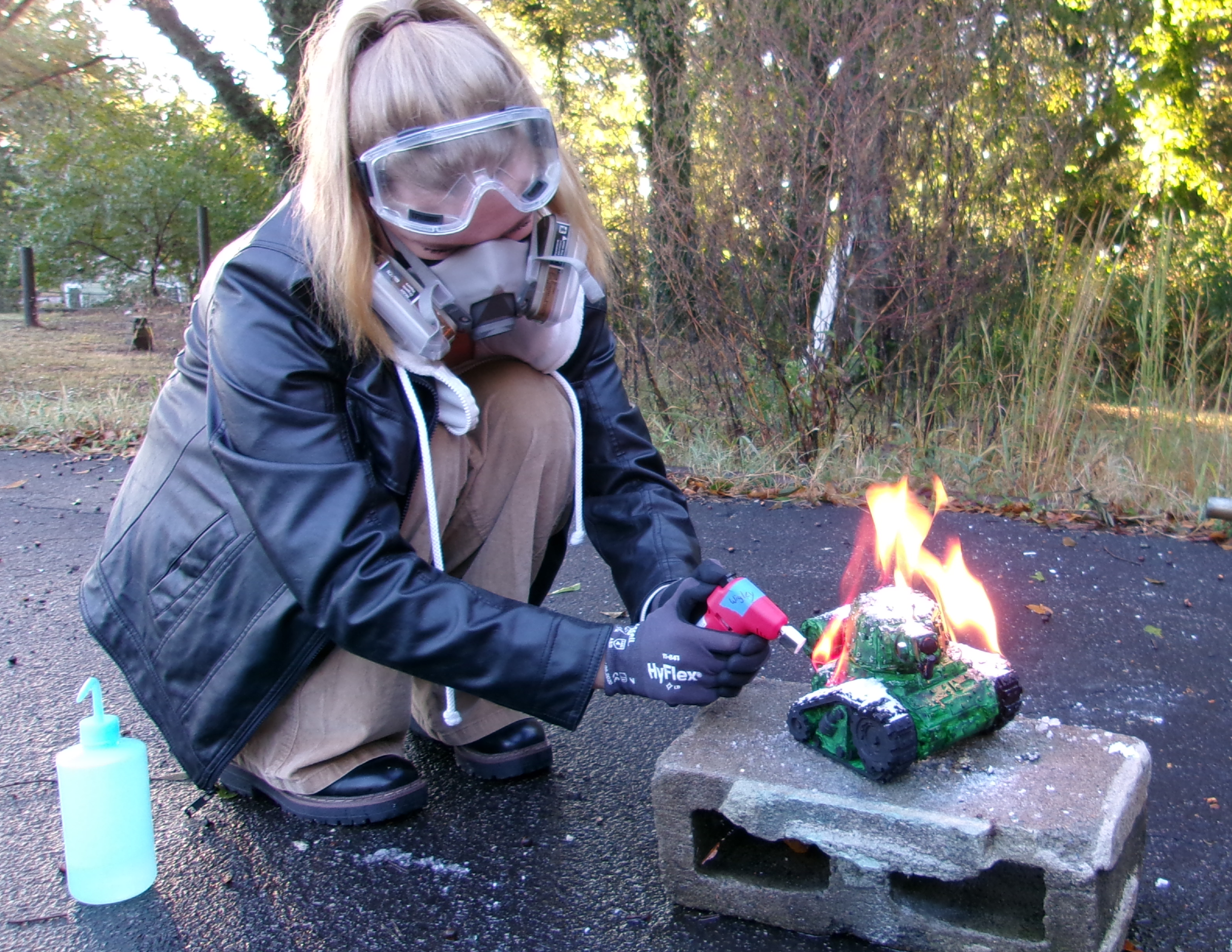 A person wearing a black leather jacket, safety goggles, a mask, and gloves is crouching while using a soldering iron on a small model tank that is on fire. The model tank is placed on a cinder block outdoors, with trees and bushes in the background.