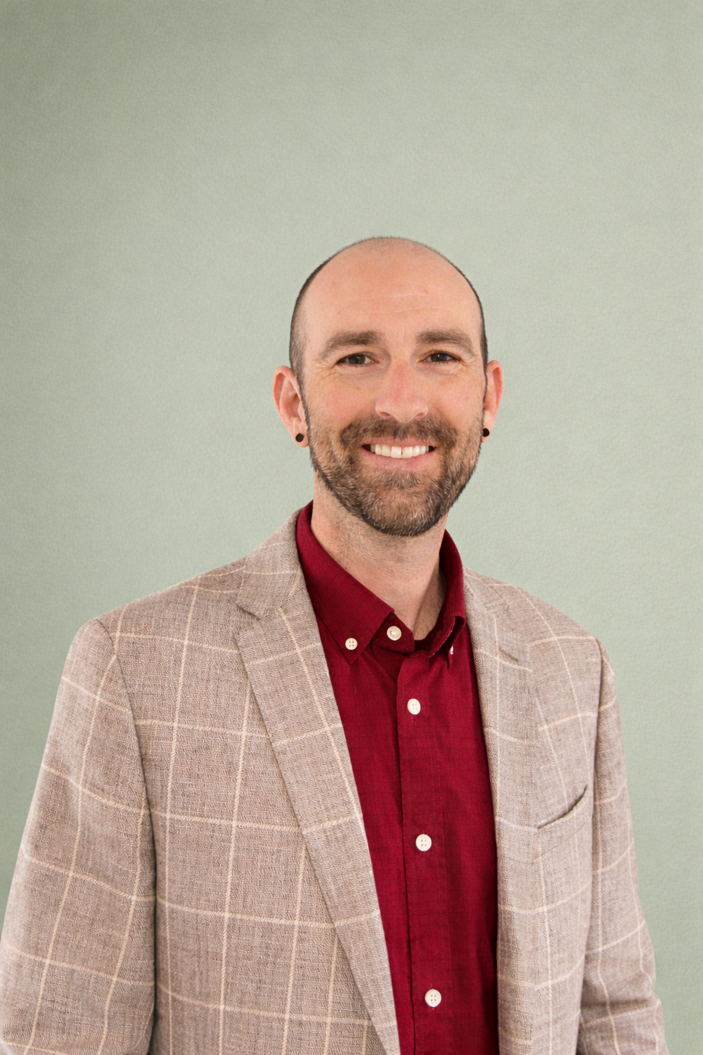 A man with a beard and earrings smiling, wearing a beige checkered blazer and a red shirt, standing against a light green background.