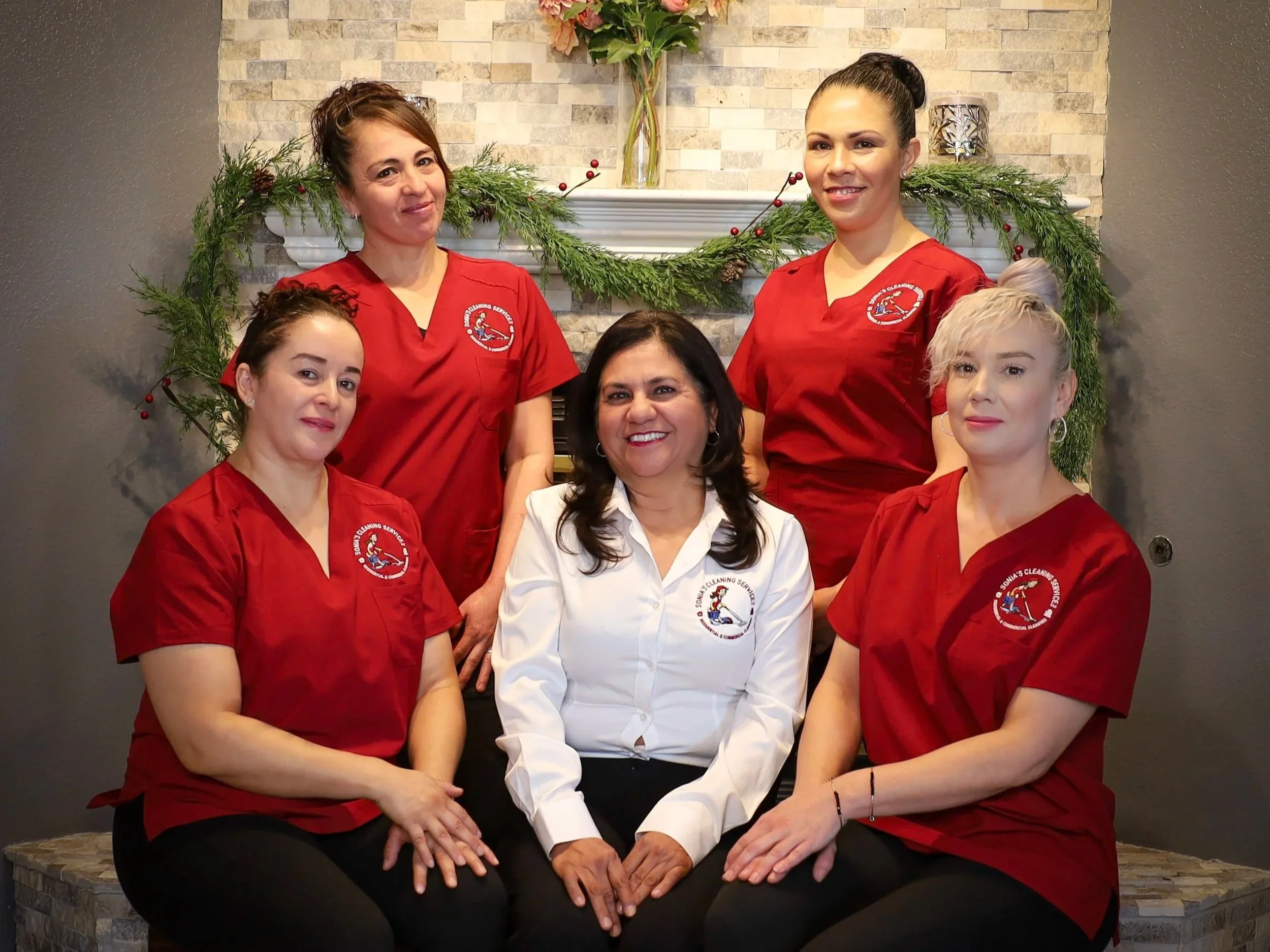 Five women, four in red medical scrubs and one in a white shirt, pose together in front of a fireplace with holiday decorations.