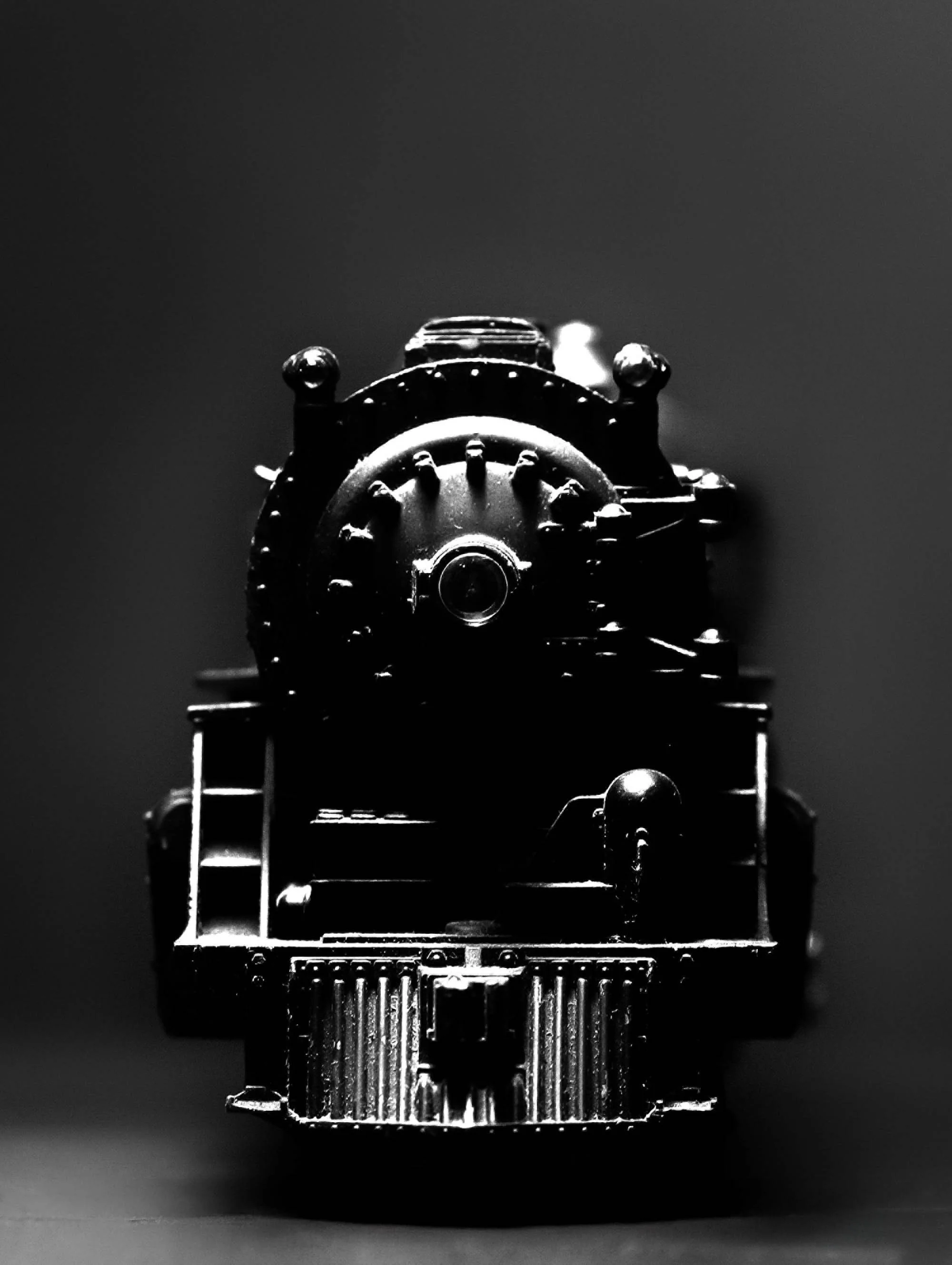 Close-up black and white photo of a vintage steam locomotive viewed from the front, with the front face of the train in sharp focus and a plain dark background.
