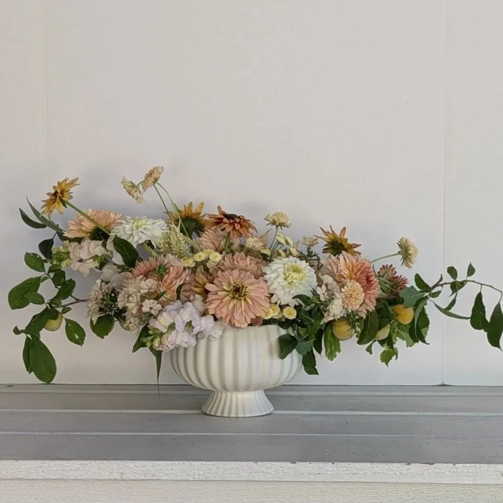 A white ceramic bowl filled with pink, white, and cream flowers along with green leaves, sitting on a gray wooden surface against a plain white wall.