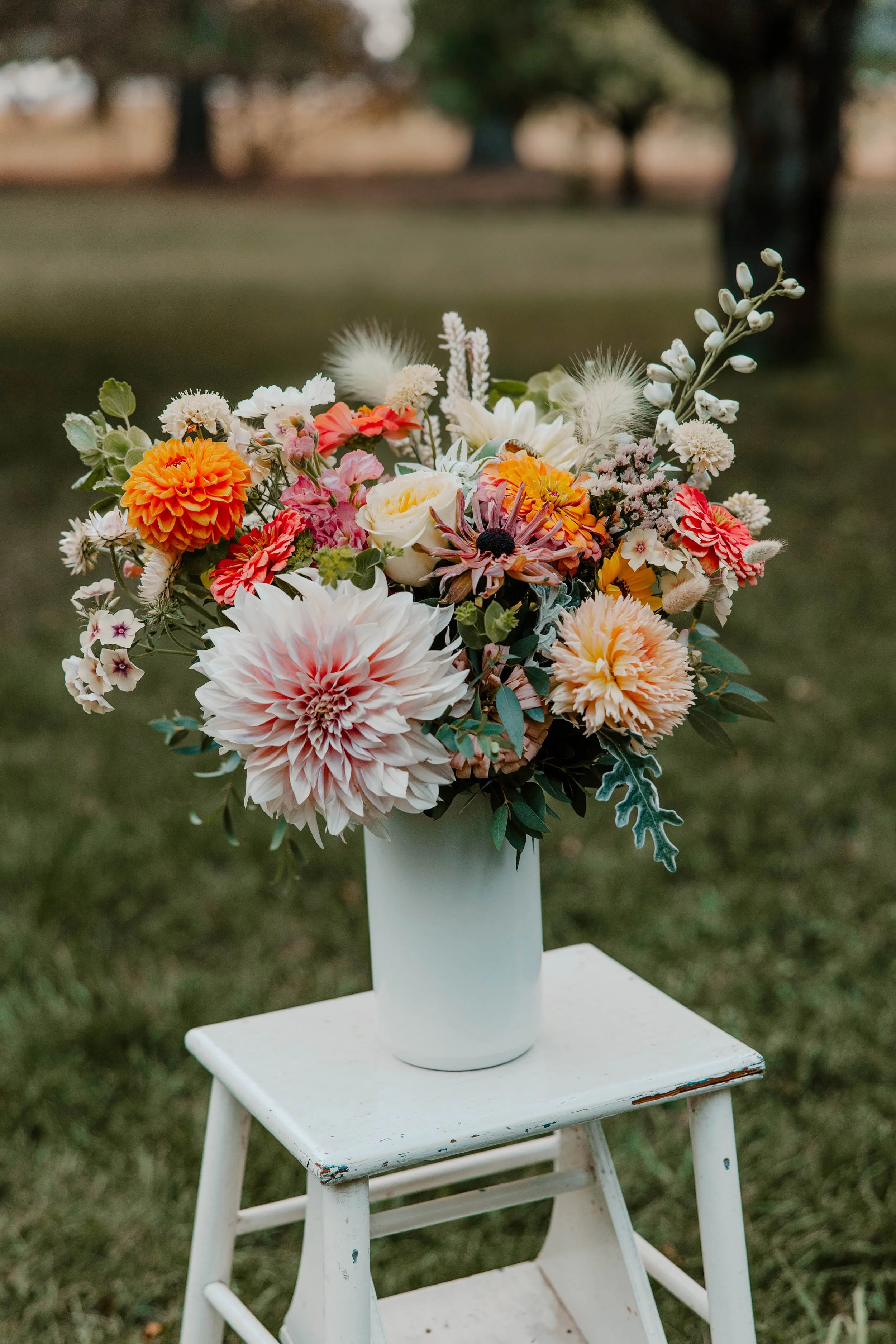 A vibrant bouquet of various flowers including dahlias, roses, and others in a white vase, placed on a small white wooden stool outdoors on grass with trees in the background.