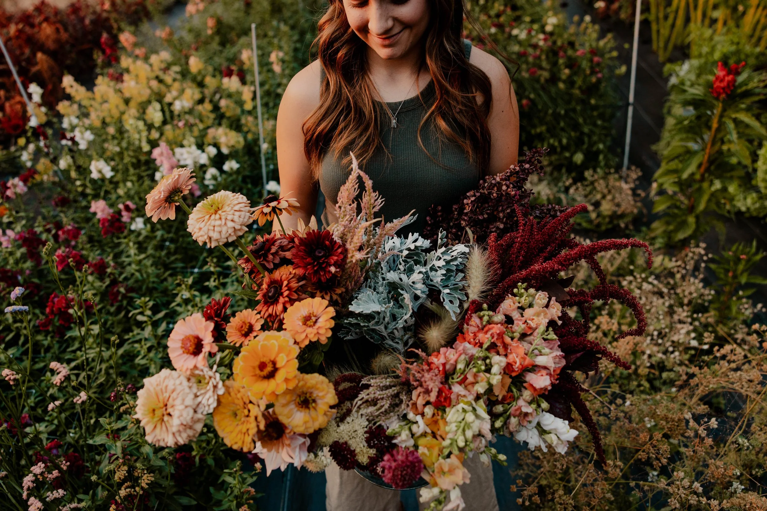 Woman holding a bucket of farm fresh flowers in pink, orangs, red, and yellow with a garden in the background.