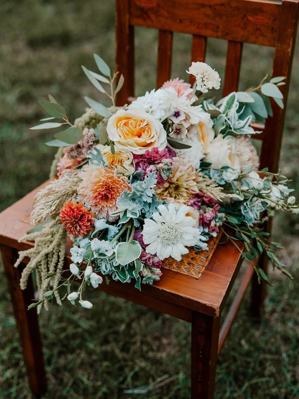 A wooden chair with a woven seat holding a large bouquet of mixed flowers, including roses, dahlias, and greenery, placed outdoors on grass.