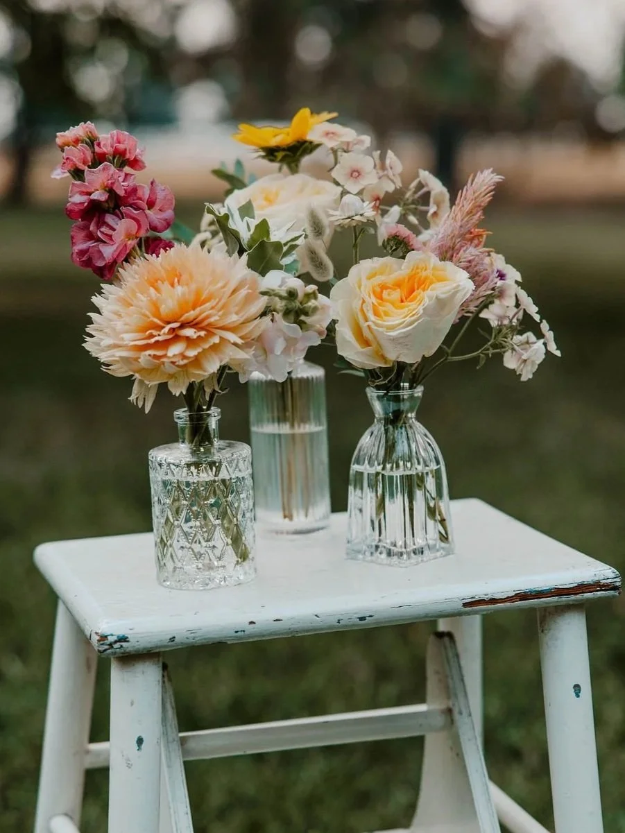 Three glass vases with fresh flowers on a white wooden stool outdoors.