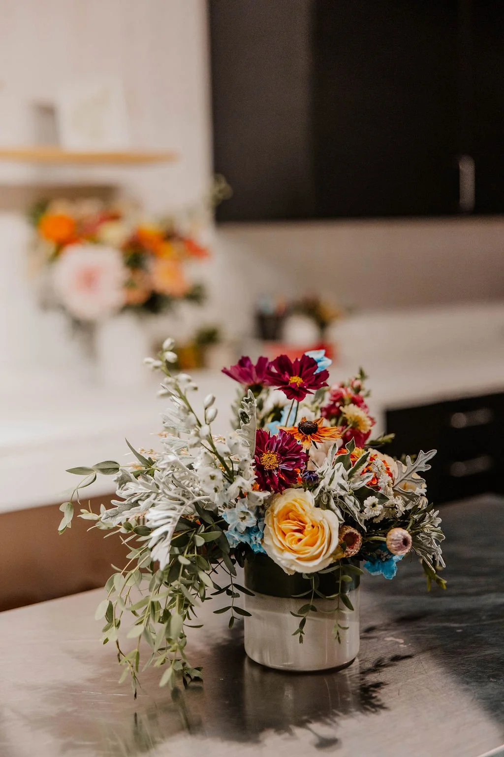 A colorful flower arrangement in a white ceramic vase on a dark wooden table, with blurred background of a kitchen with flowers and decorations.