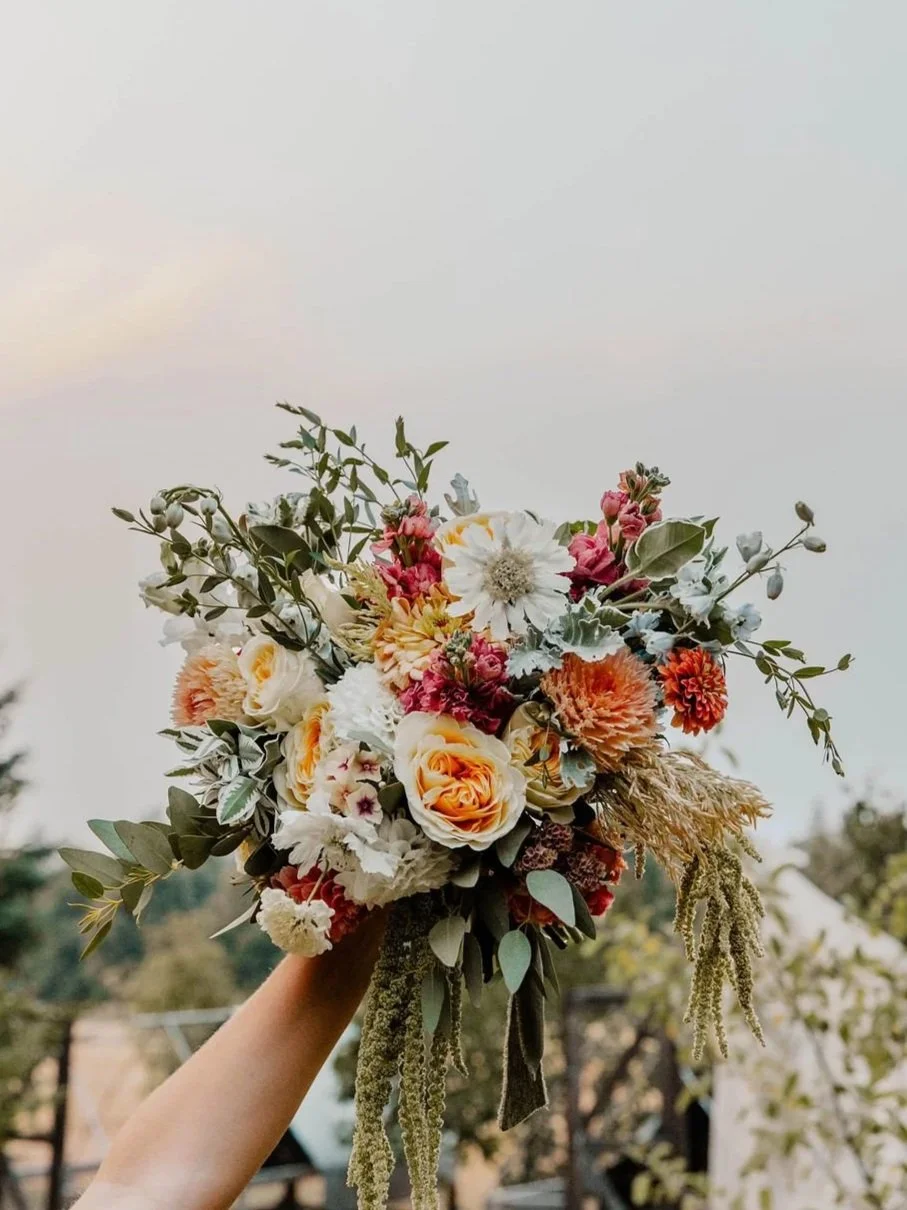 A person holding a bouquet of mixed flowers including roses, dahlias , and foliage outdoors against a cloudy sky.