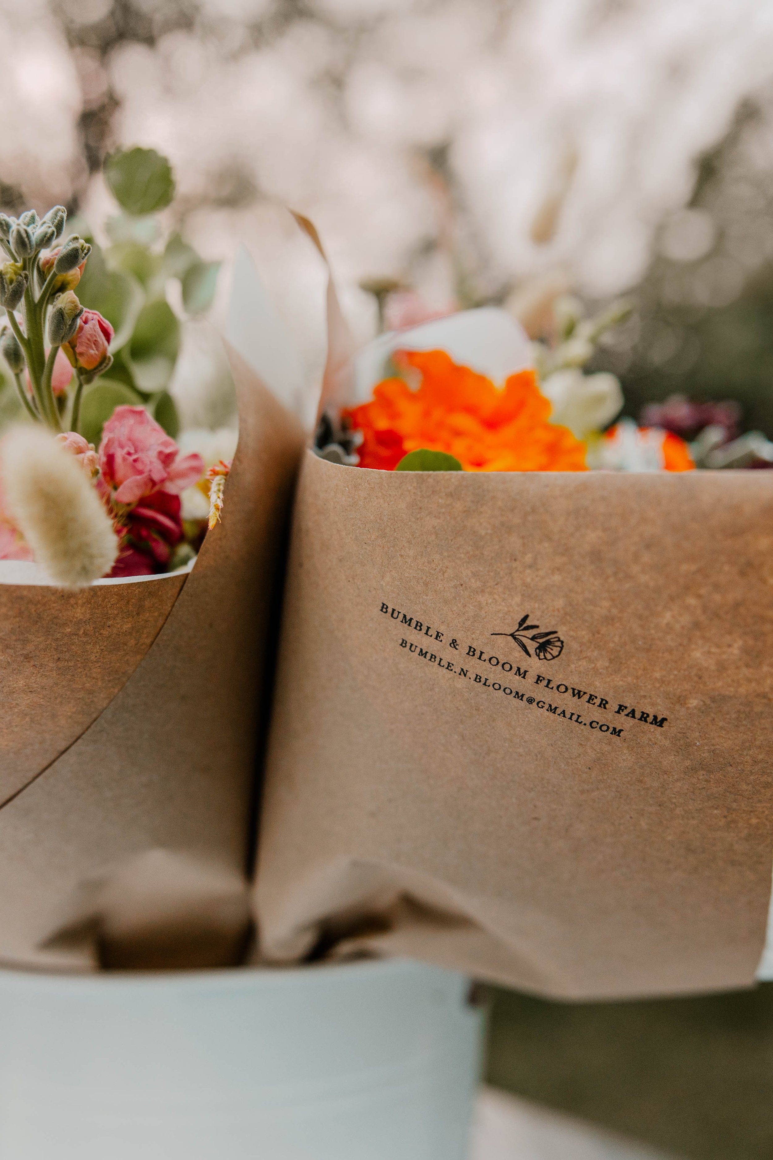 Close-up of a bouquet of flowers wrapped in brown paper, with text printed on the paper that reads 'Bumble & Bloom Flower Farm' along with the farm's email address.