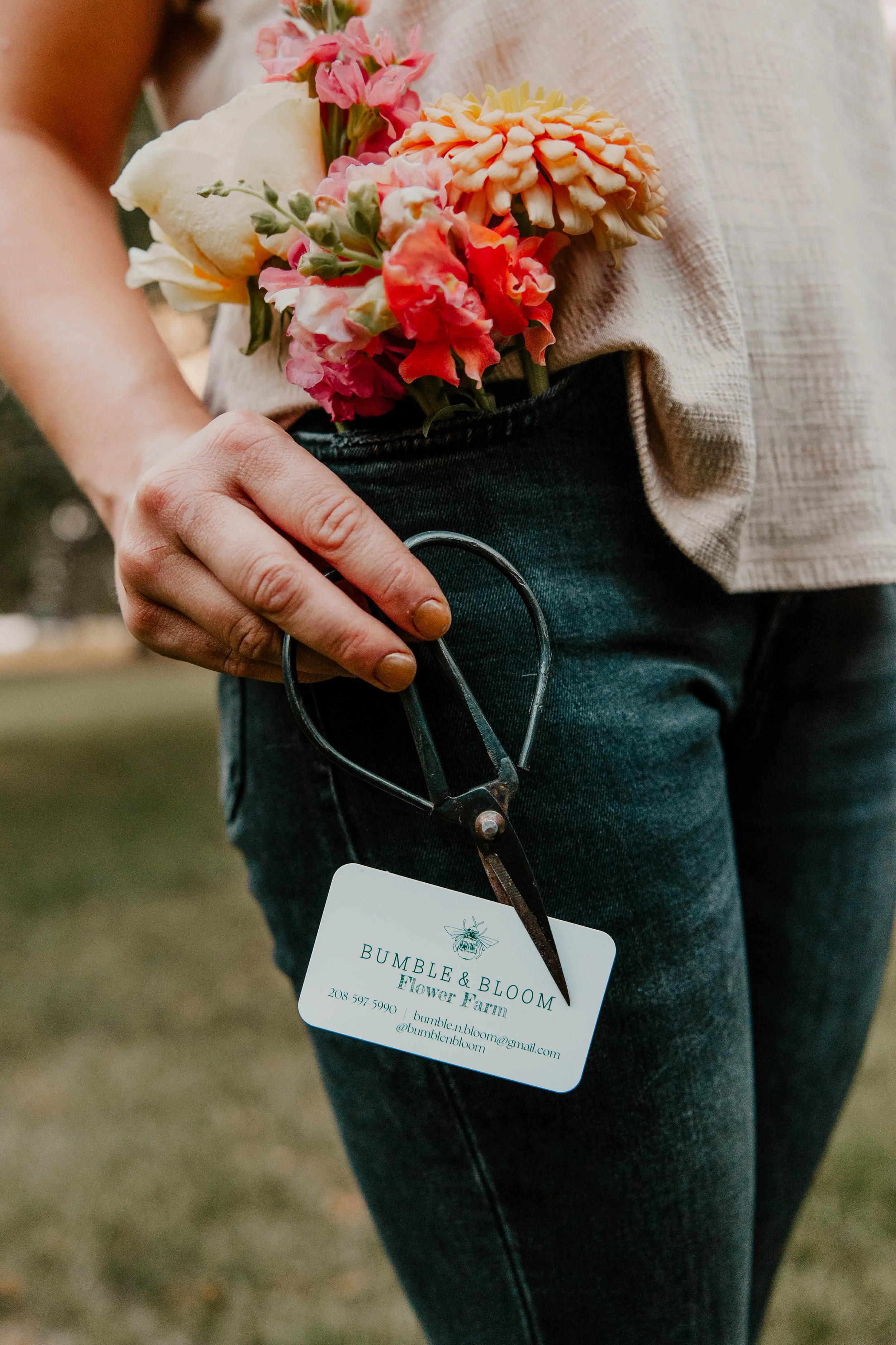 Person with black pants standing outdoors holding a pair of black scissors next to their pocket with orange and yellow flowers in the pocket.