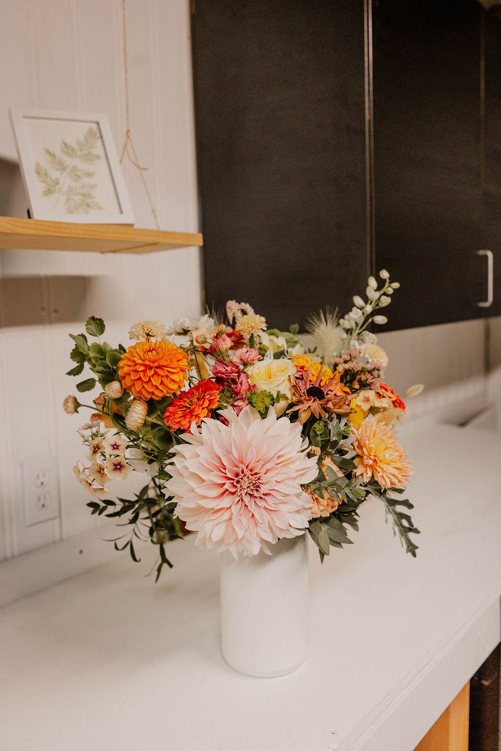 A bouquet of colorful flowers in a white vase on a white surface, with a black cabinet and a framed botanical print in the background.