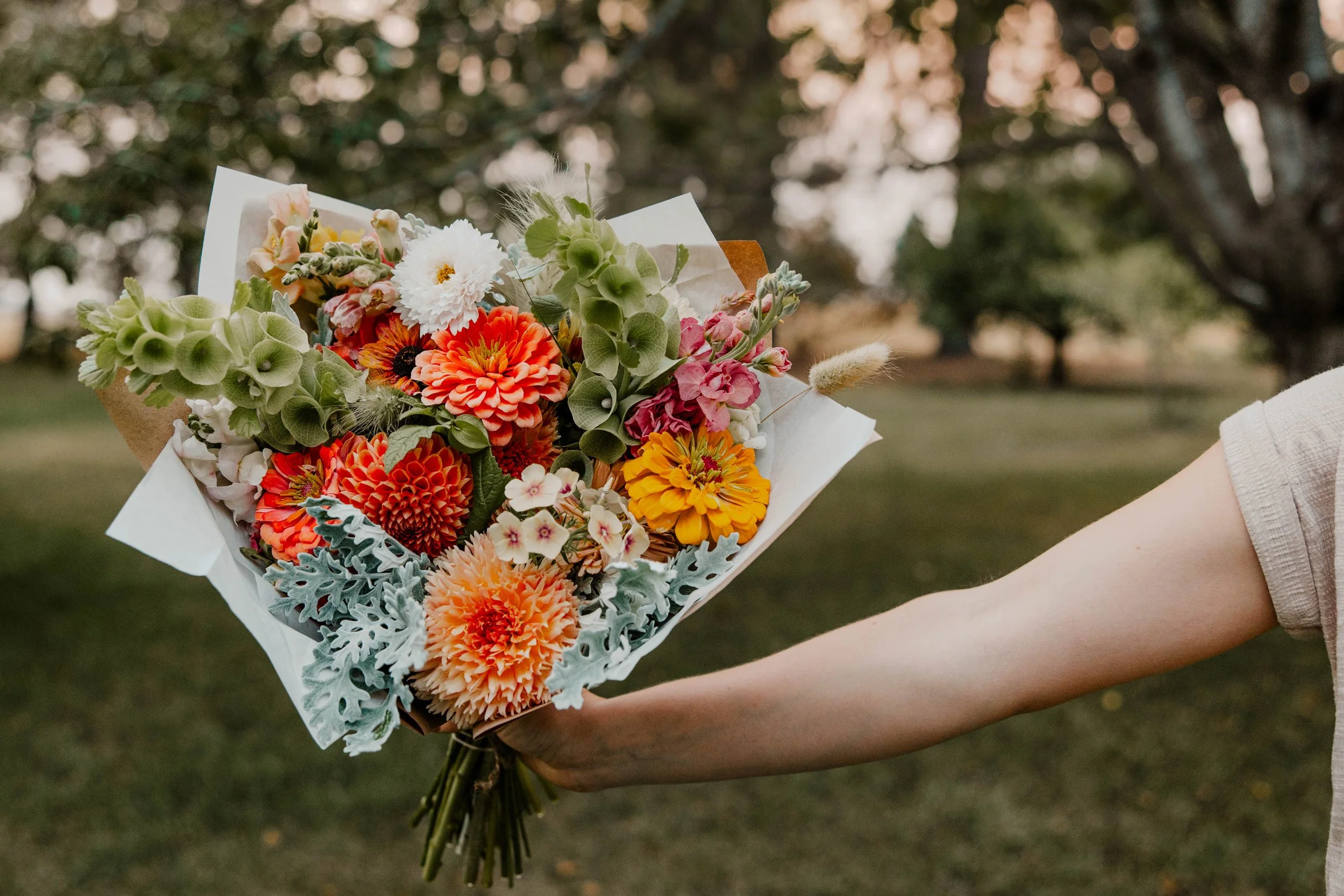 A person's hand holding a colorful bouquet of flowers including orange dahlias, pink stock, and green bells of Ireland with blurred green trees in the background.
