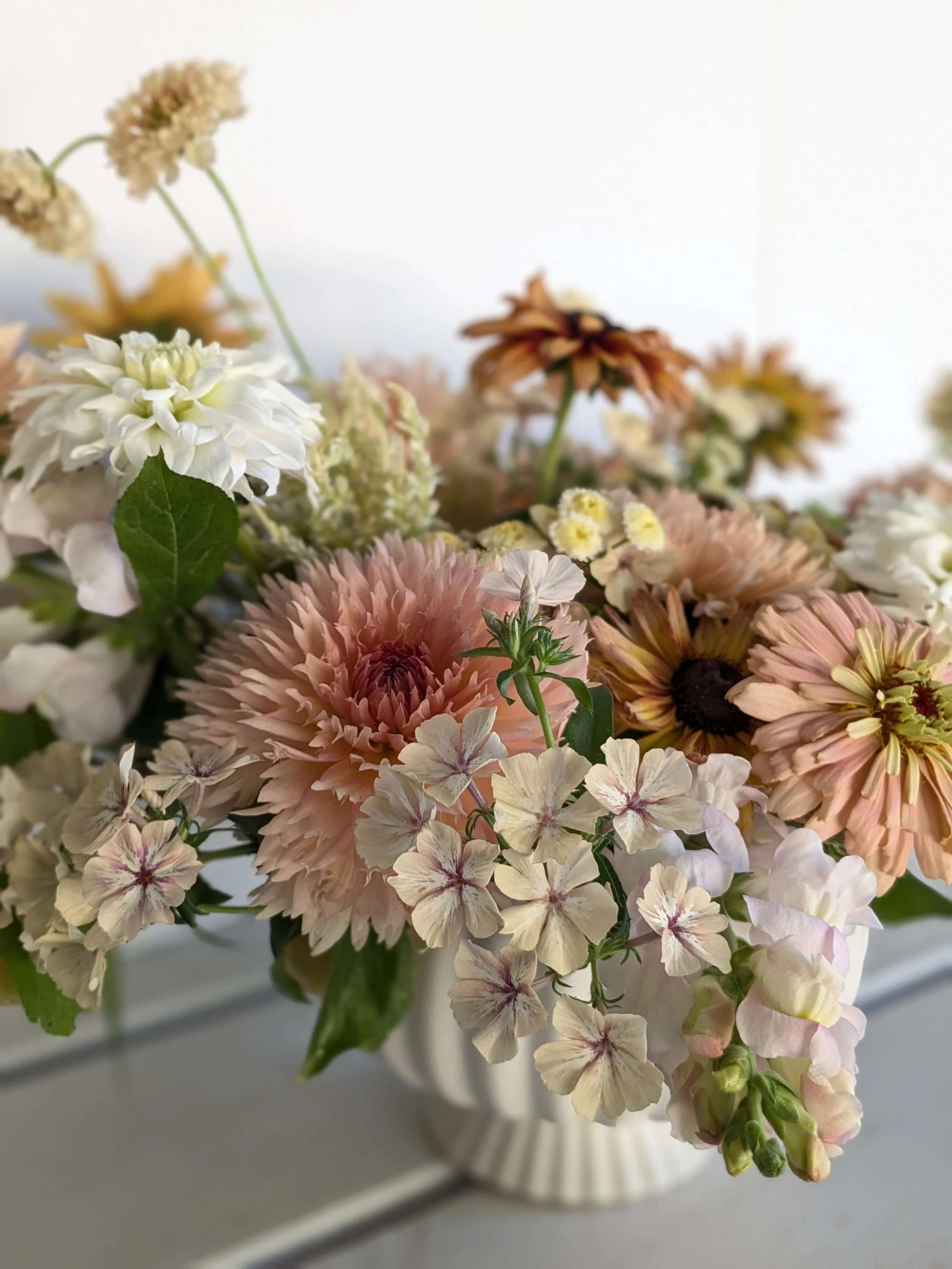 A bouquet of various pastel-colored flowers in a white ceramic vase against a plain light background.