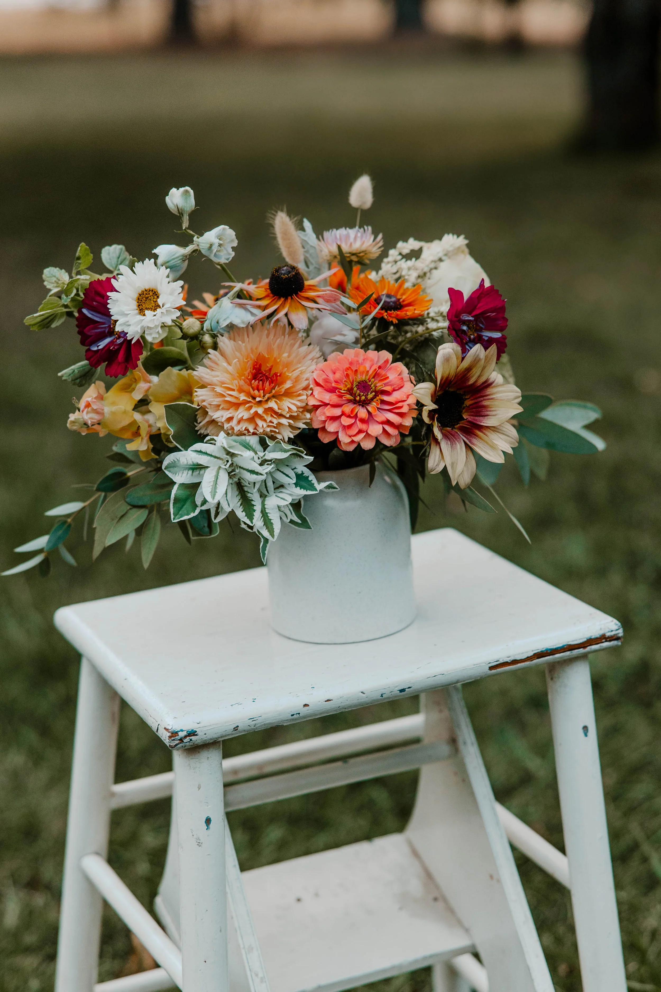 A bouquet of colorful flowers in a white vase placed on a small, white, distressed wooden table outdoors.