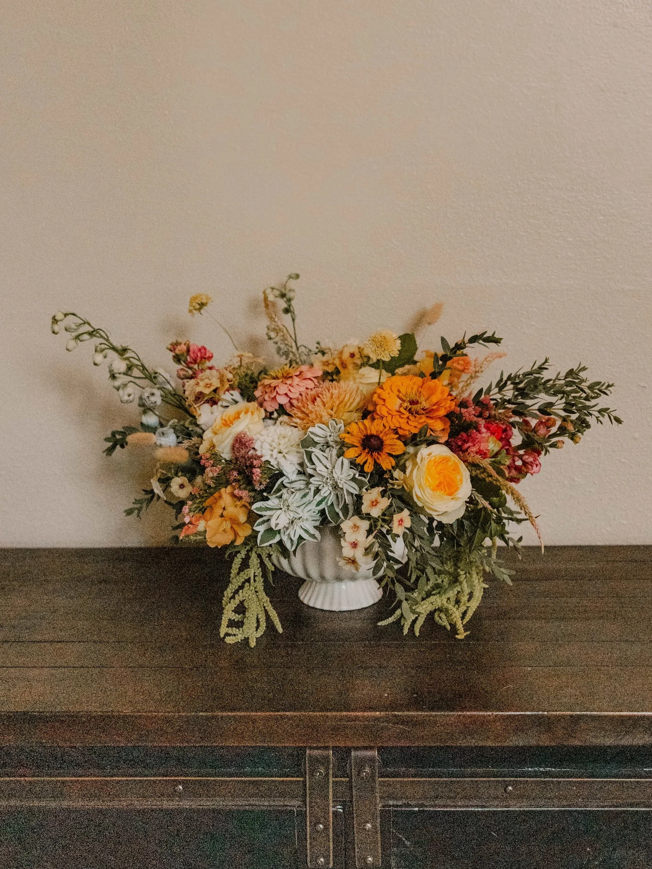 A floral arrangement with orange, pink, yellow, and white flowers in a white ceramic vase on a dark wooden surface against a plain beige wall.
