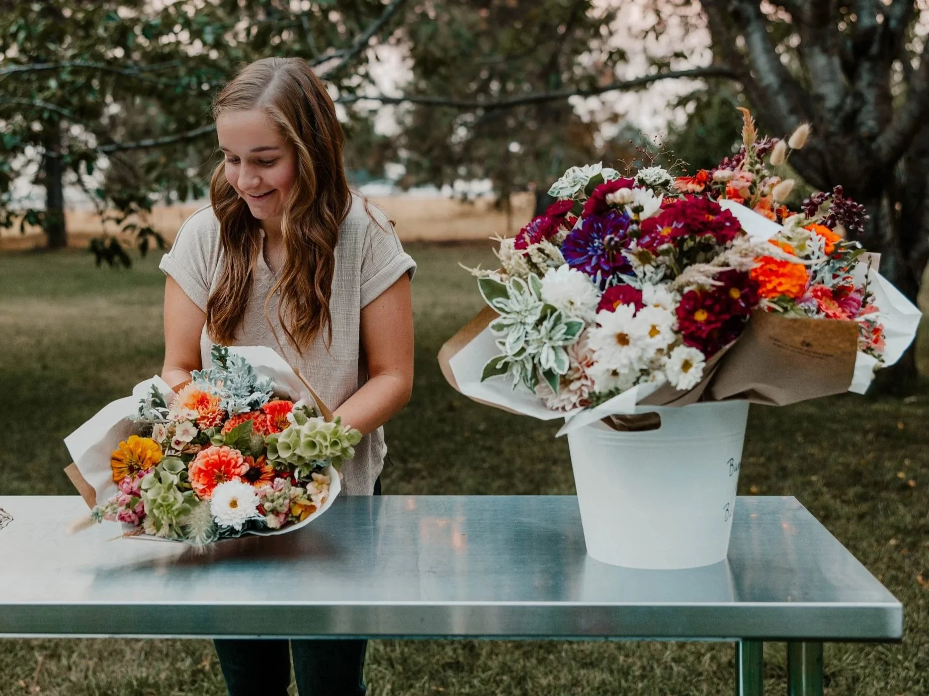 A young woman arranging a colorful bouquet of flowers on a metal table outdoors, with another large bouquet on the table and trees in the background.