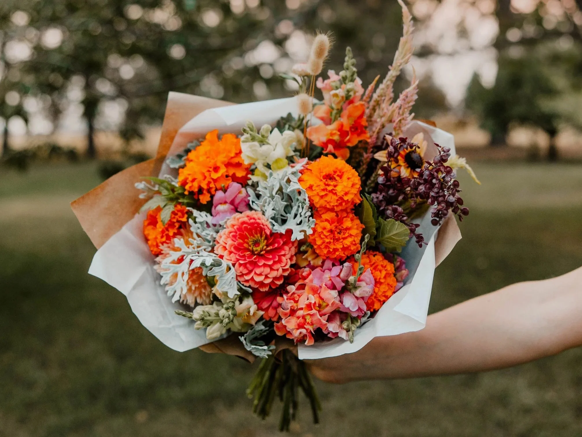 A person holding a colorful bouquet of orange, pink, white, and yellow flowers wrapped in brown and white paper, outdoors with trees in the background.