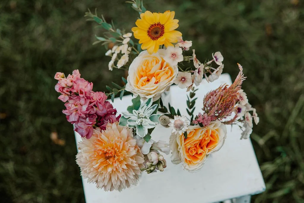 A bouquet of various colorful flowers including yellow daisies, pink snapdragons, peach roses, and white blossoms, arranged on a white surface outdoors.