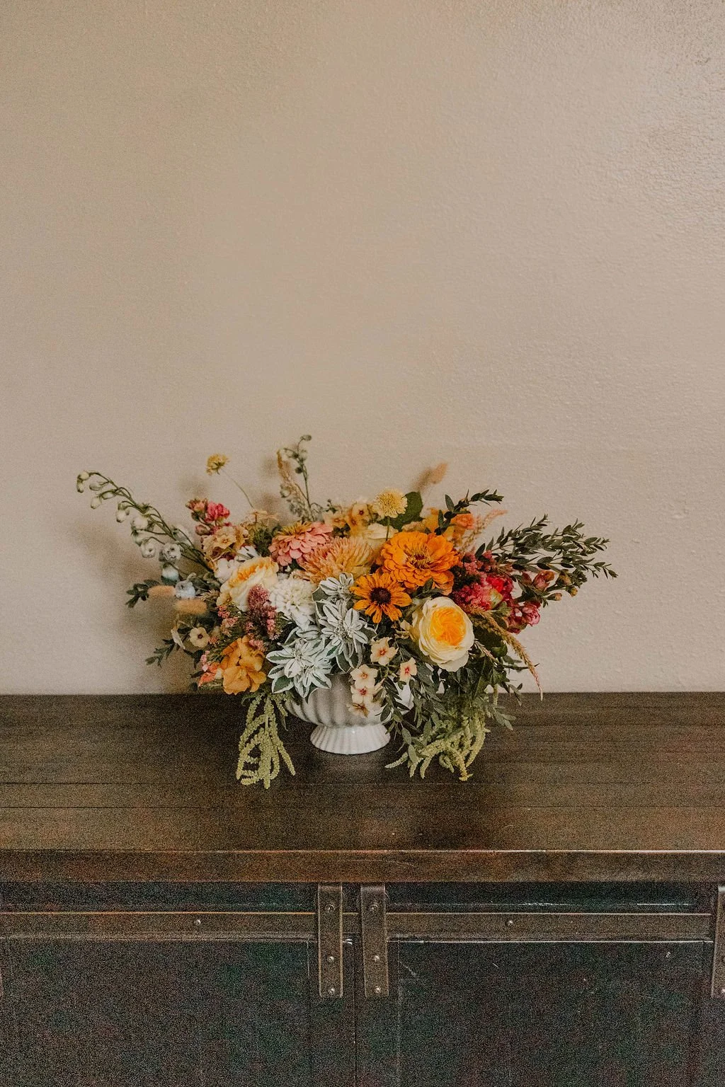 A colorful bouquet of flowers in a white vase on a dark wooden surface against a plain wall.