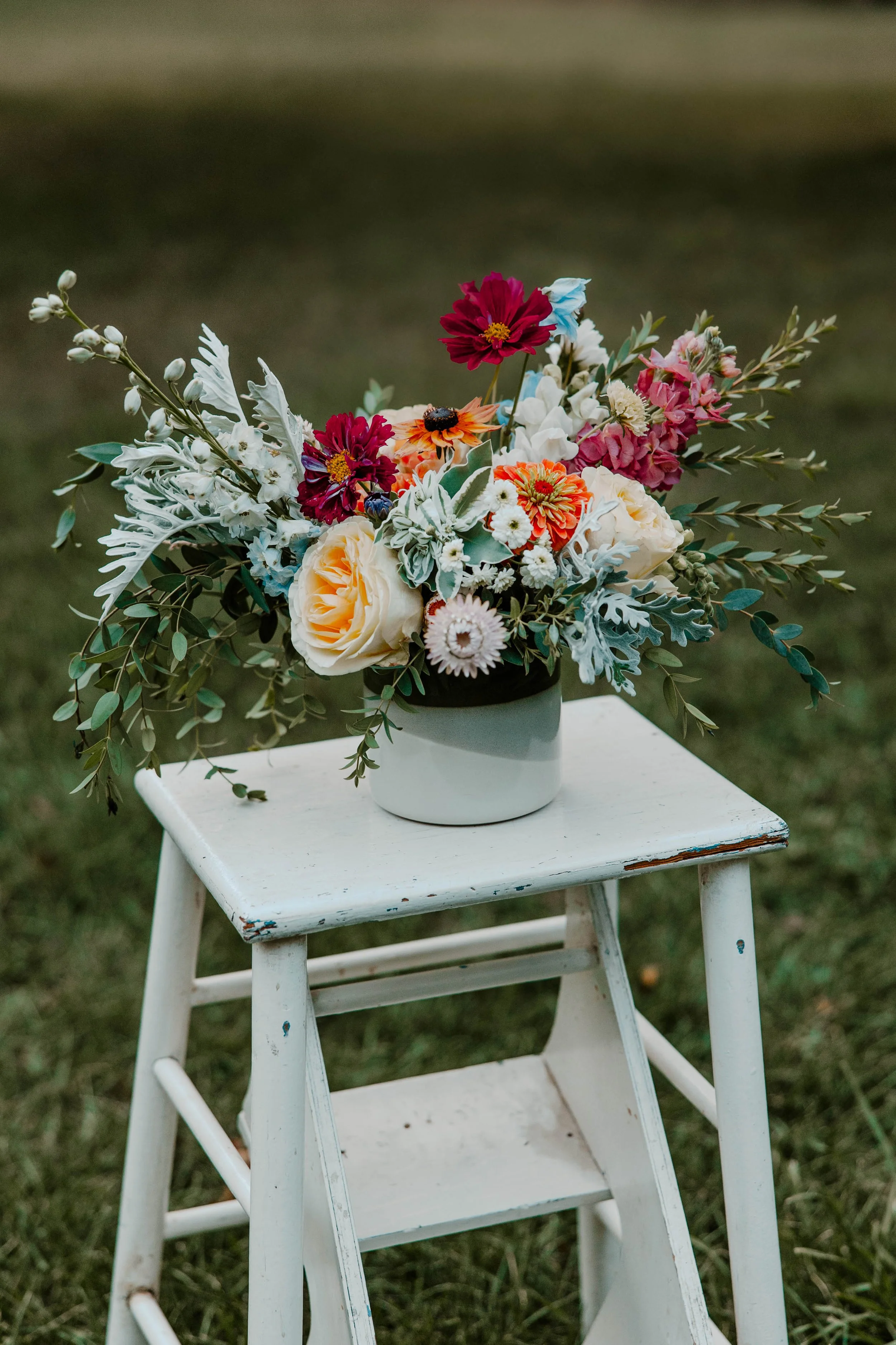 A bouquet of mixed flowers, including roses, daisies, and other blooms, arranged in a black and white vase, placed on a white wooden stool outdoors.